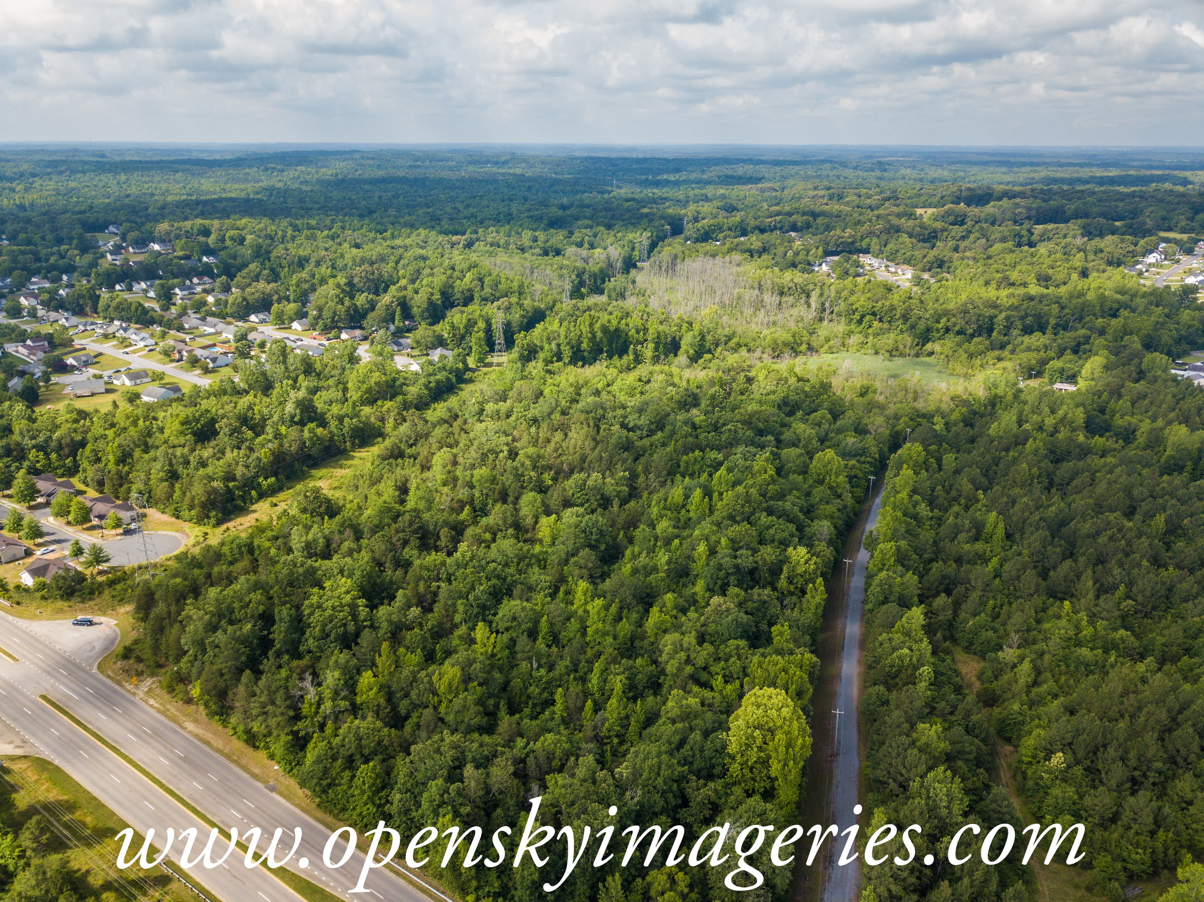 Aerial drone photograph of a large wooded tract surrounded by residential neighborhoods, with a multilane roadway along one side, a narrow access road through the trees, and forest extending toward the horizon.