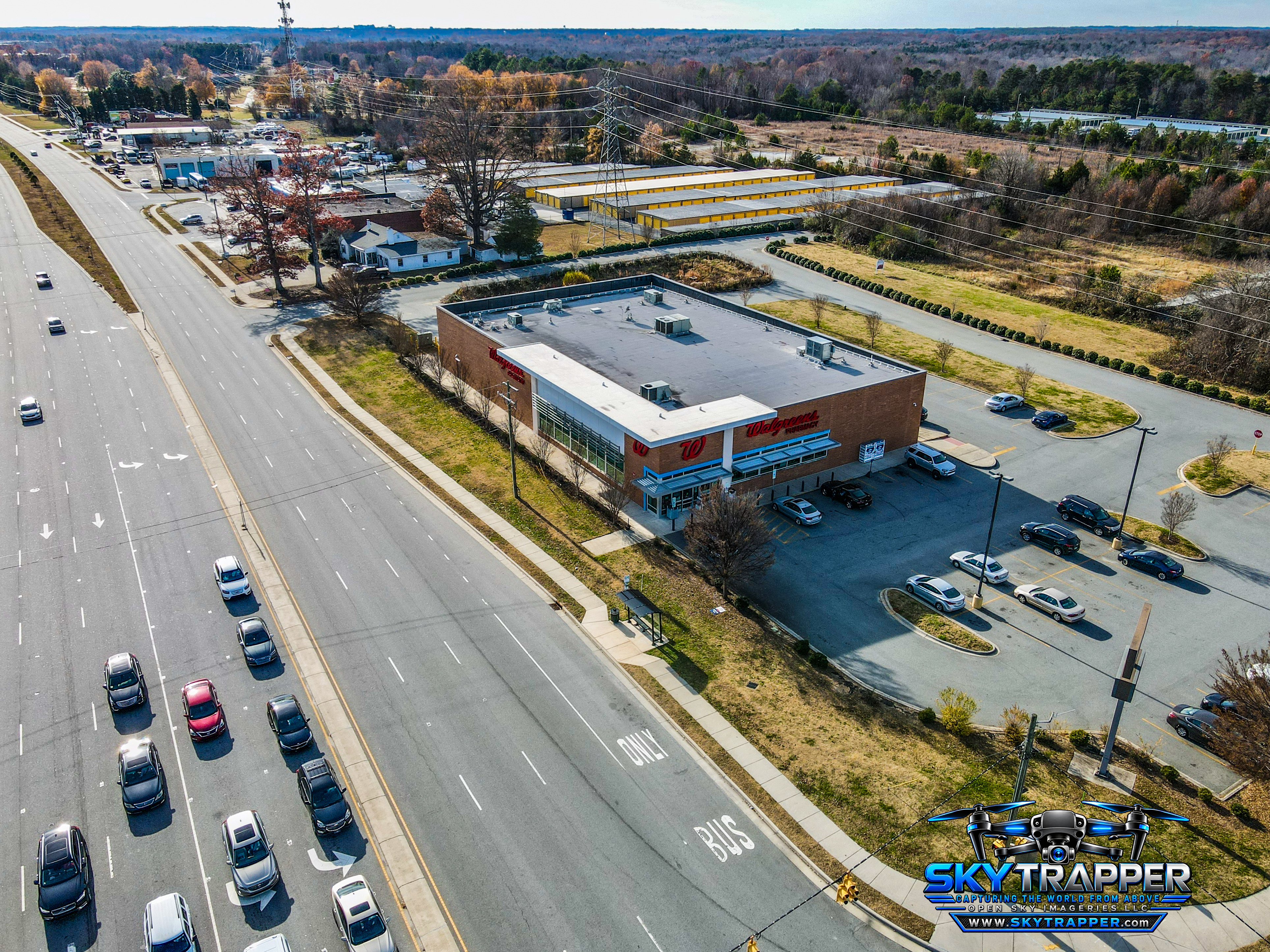 Aerial photograph of a Walgreens retail pharmacy with a flat-roofed brick building, parking lot, adjacent multilane roadway, nearby commercial buildings, and surrounding wooded areas.