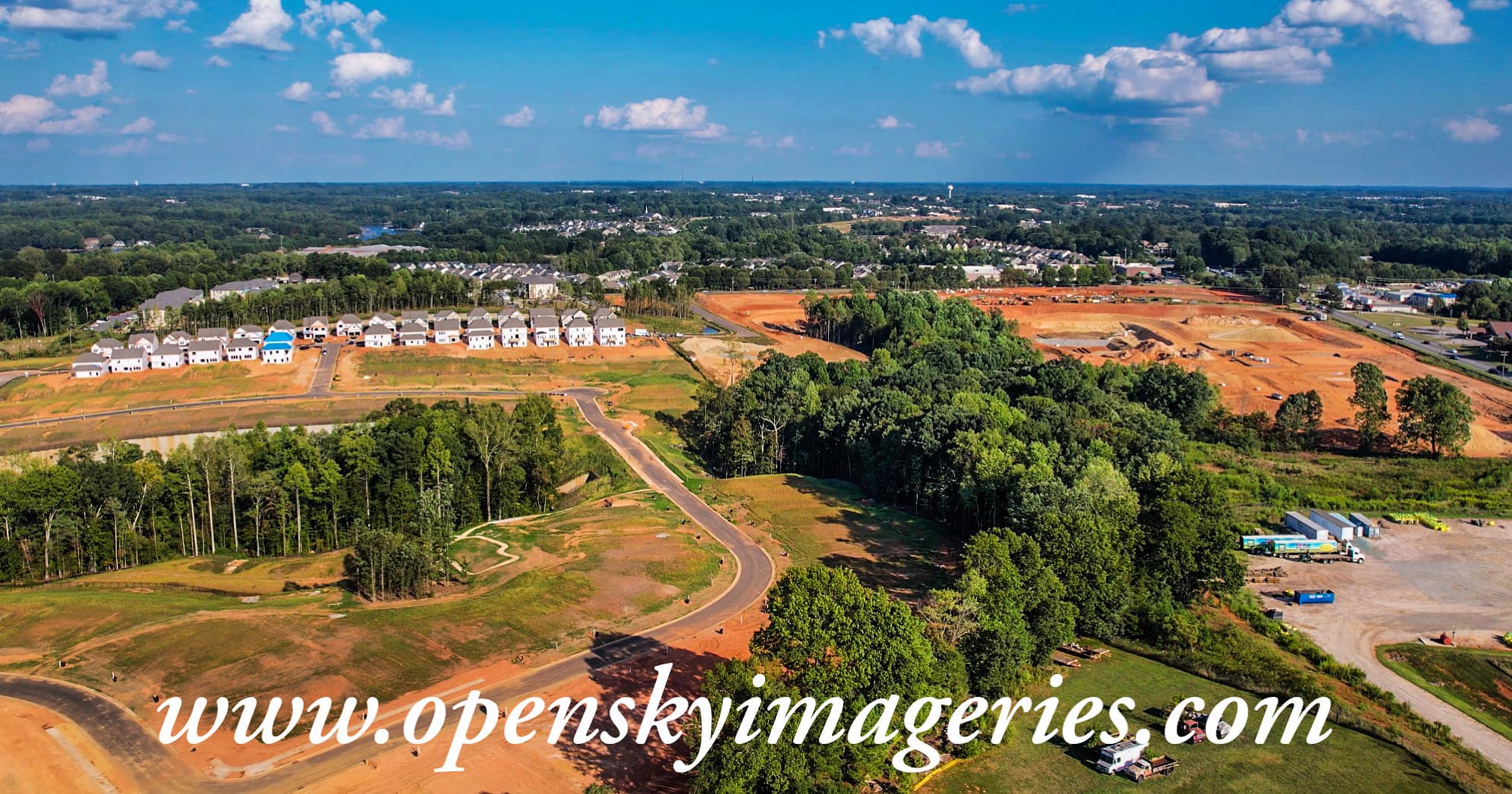 Wide aerial drone view of a large residential development featuring newly constructed houses, graded construction sites, winding paved roads, earthmoving areas, wooded sections, and surrounding suburban neighborhoods.