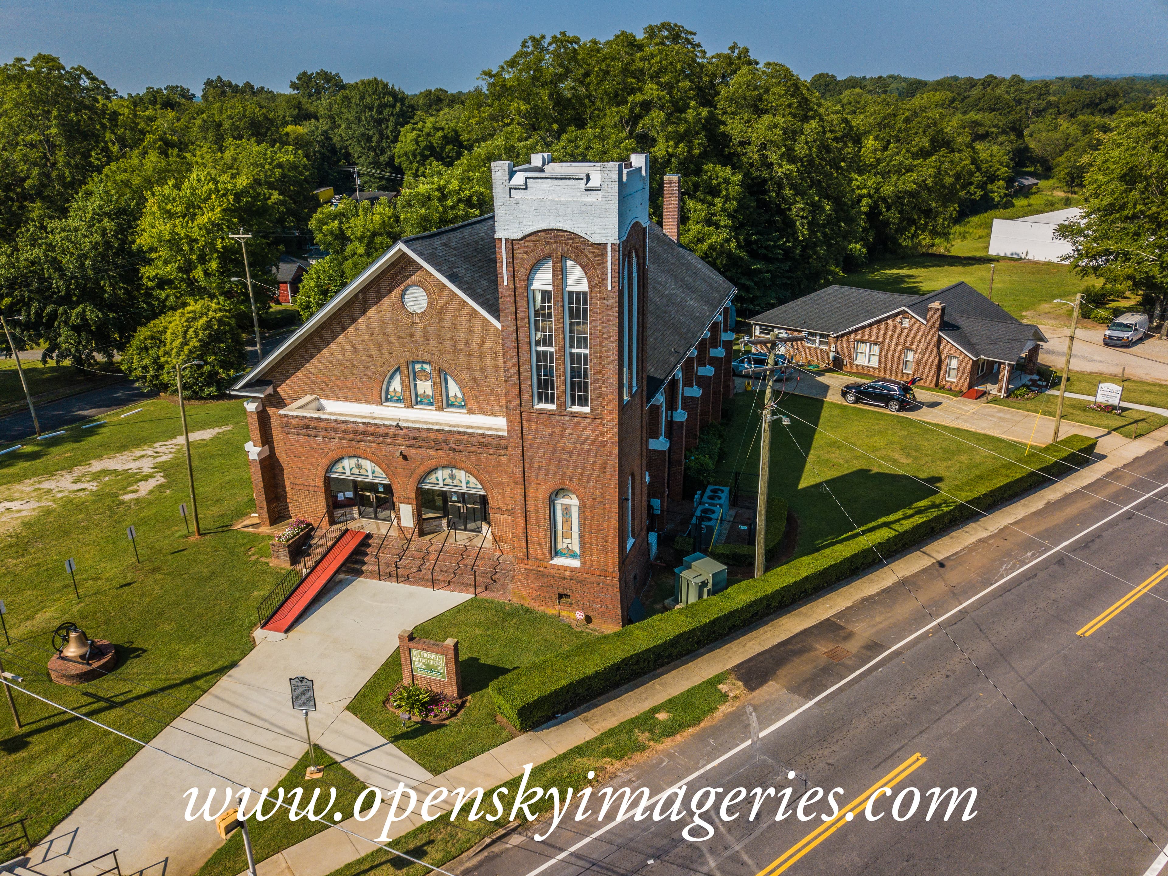 Aerial drone photograph of a historic-style red brick church with a prominent square bell tower, arched stained-glass windows, accessible entrance ramp, landscaped grounds, and adjacent roadway.