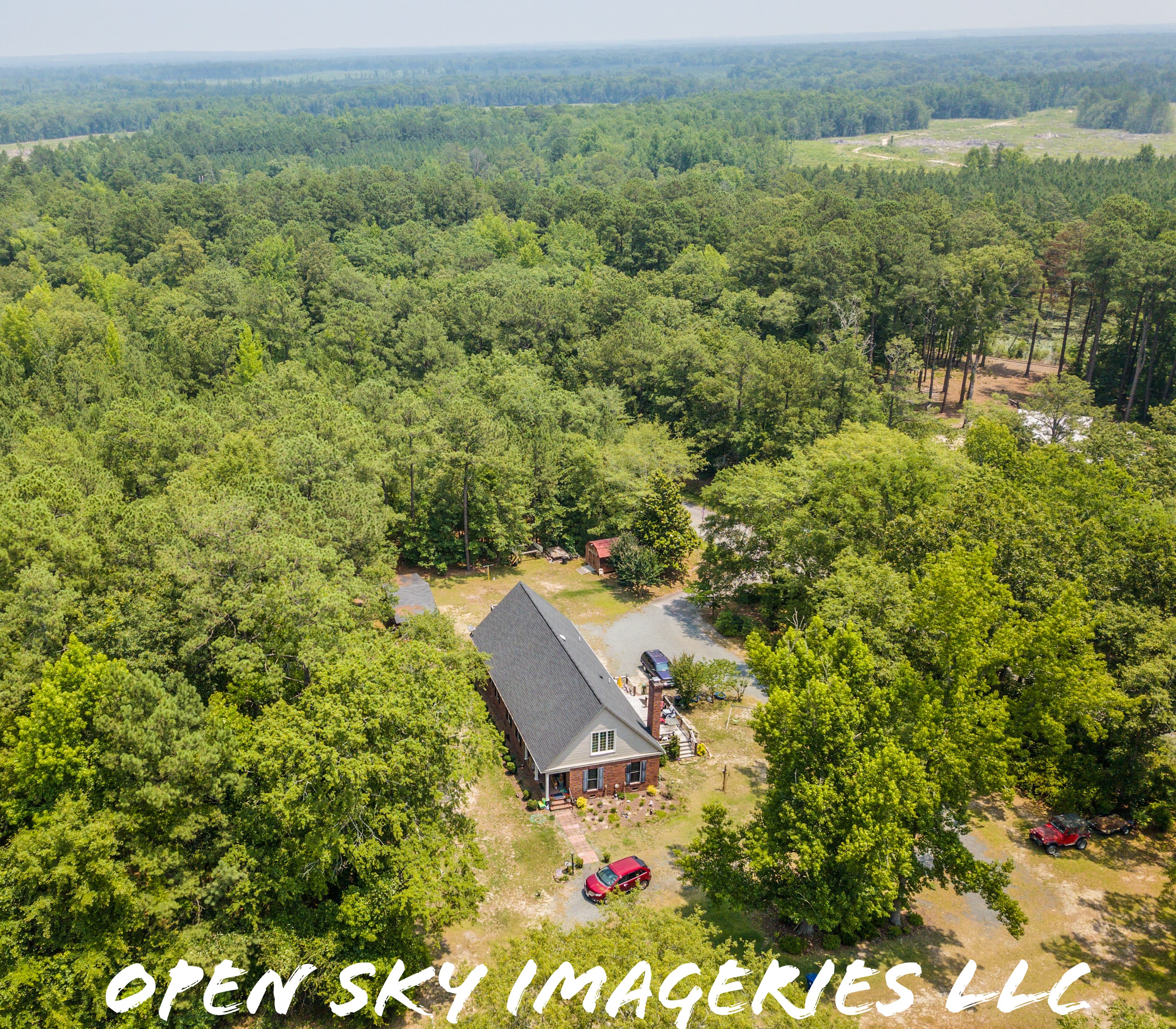 Aerial drone photograph of a secluded brick residence with a steep dark roof, surrounded by dense woodland, mature trees, a gravel driveway, parked vehicles, and a small outbuilding.