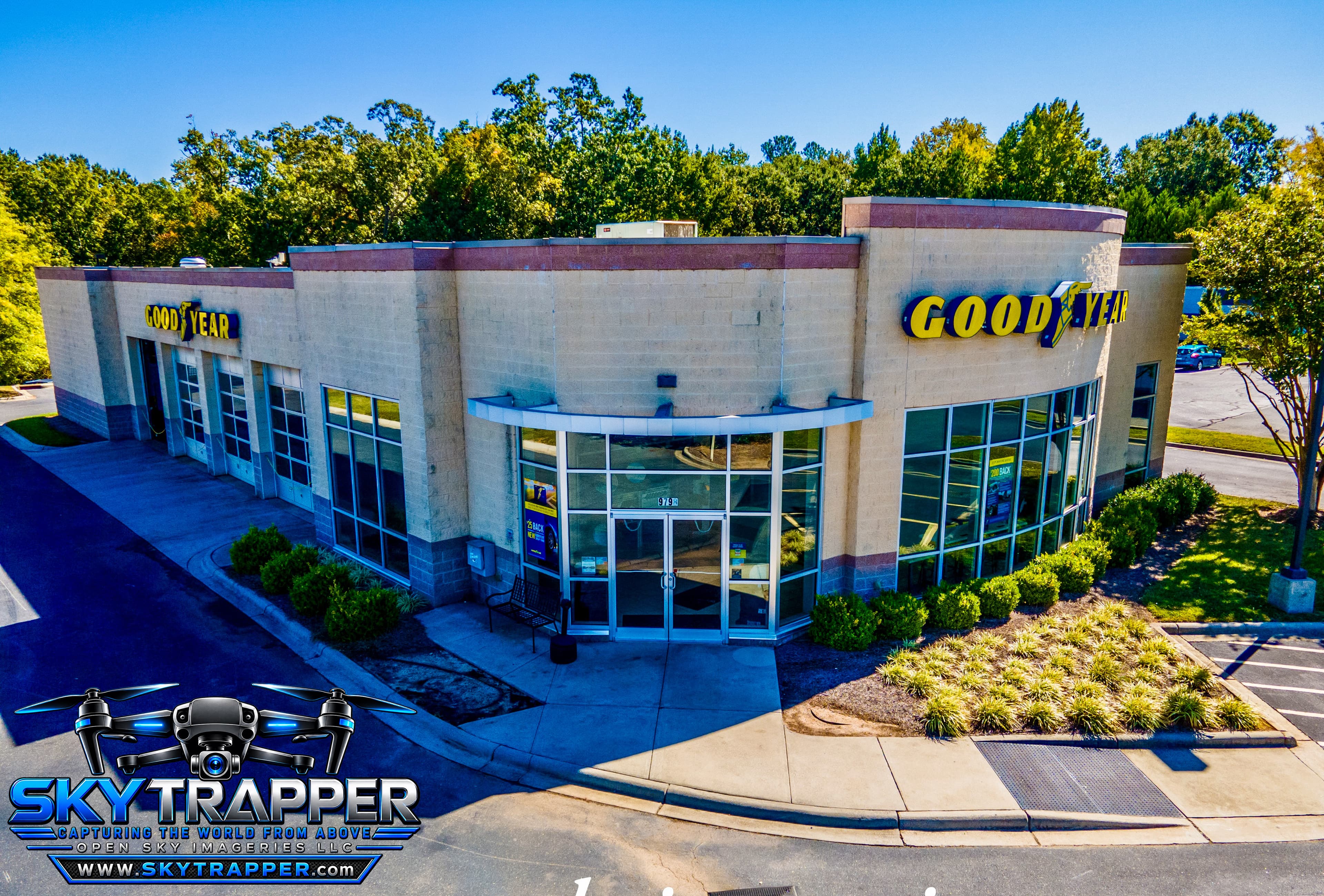 Aerial drone photograph of a Goodyear automotive service center featuring branded storefront signage, multiple vehicle service bays, a glass-front entrance, landscaped grounds, and surrounding parking areas.