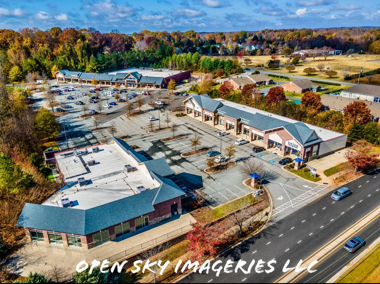 Aerial drone photograph of a suburban retail shopping center featuring multiple brick storefront buildings, expansive parking areas, landscaped islands, roadway access, and surrounding residential neighborhoods with colorful autumn foliage.