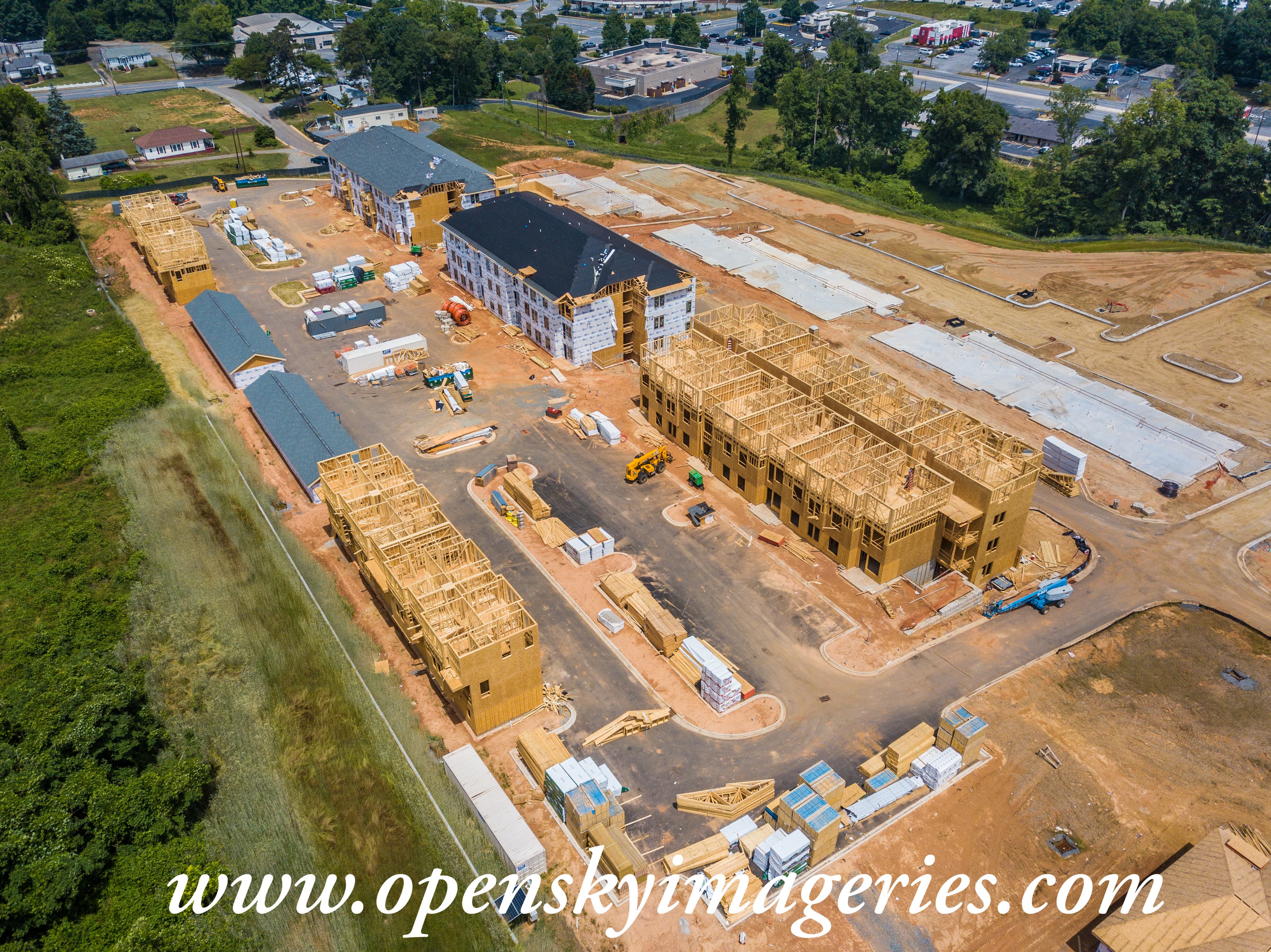 Aerial drone photograph of a multifamily residential construction site showing apartment buildings at various stages of completion, exposed wood framing, roof installation, concrete foundations, construction equipment, and staged building materials.