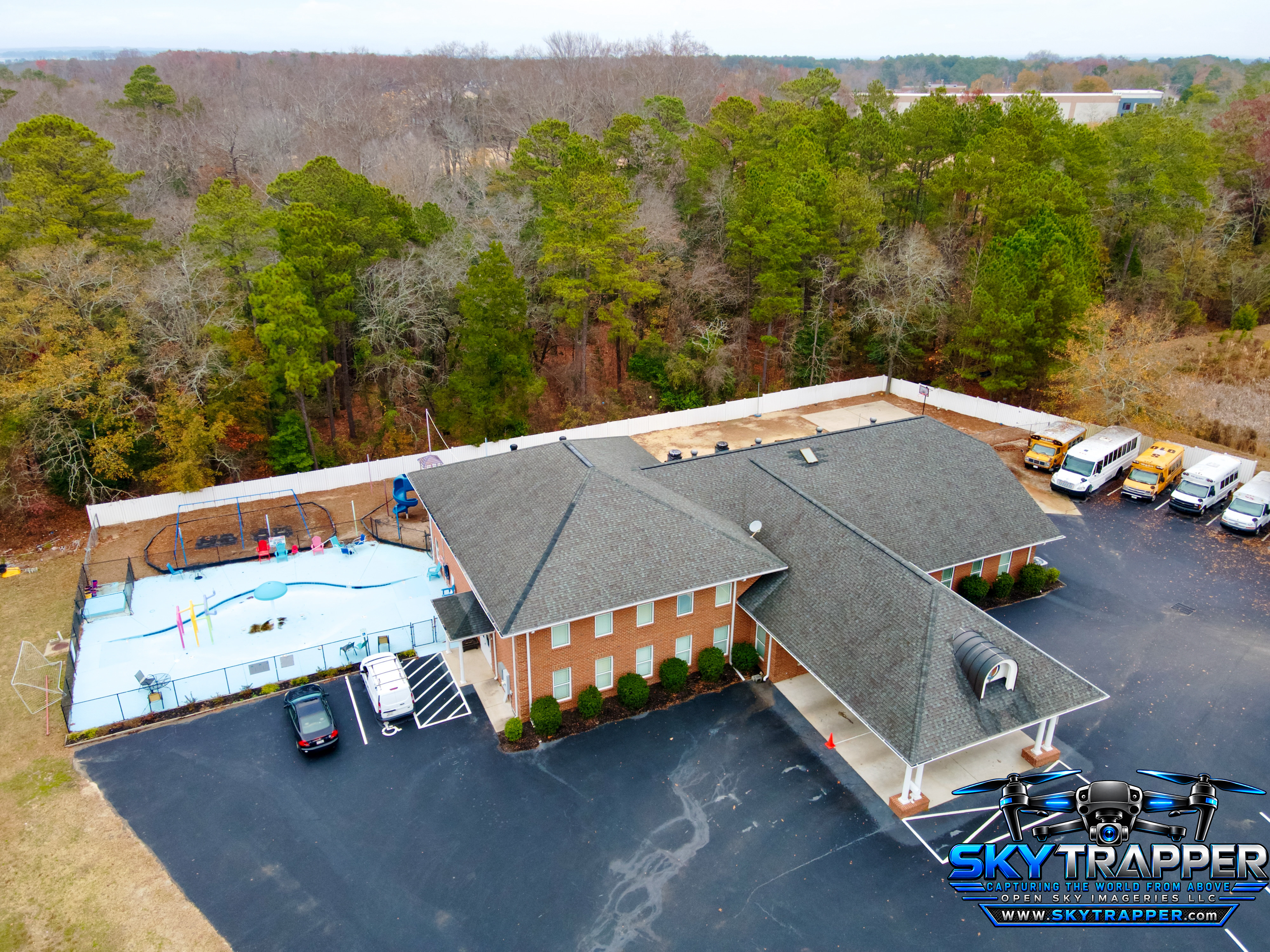 Elevated drone photograph of a large brick institutional building with a gray shingle roof, covered entrance, parking areas, several buses and vans, fenced outdoor play area, and surrounding woodland.