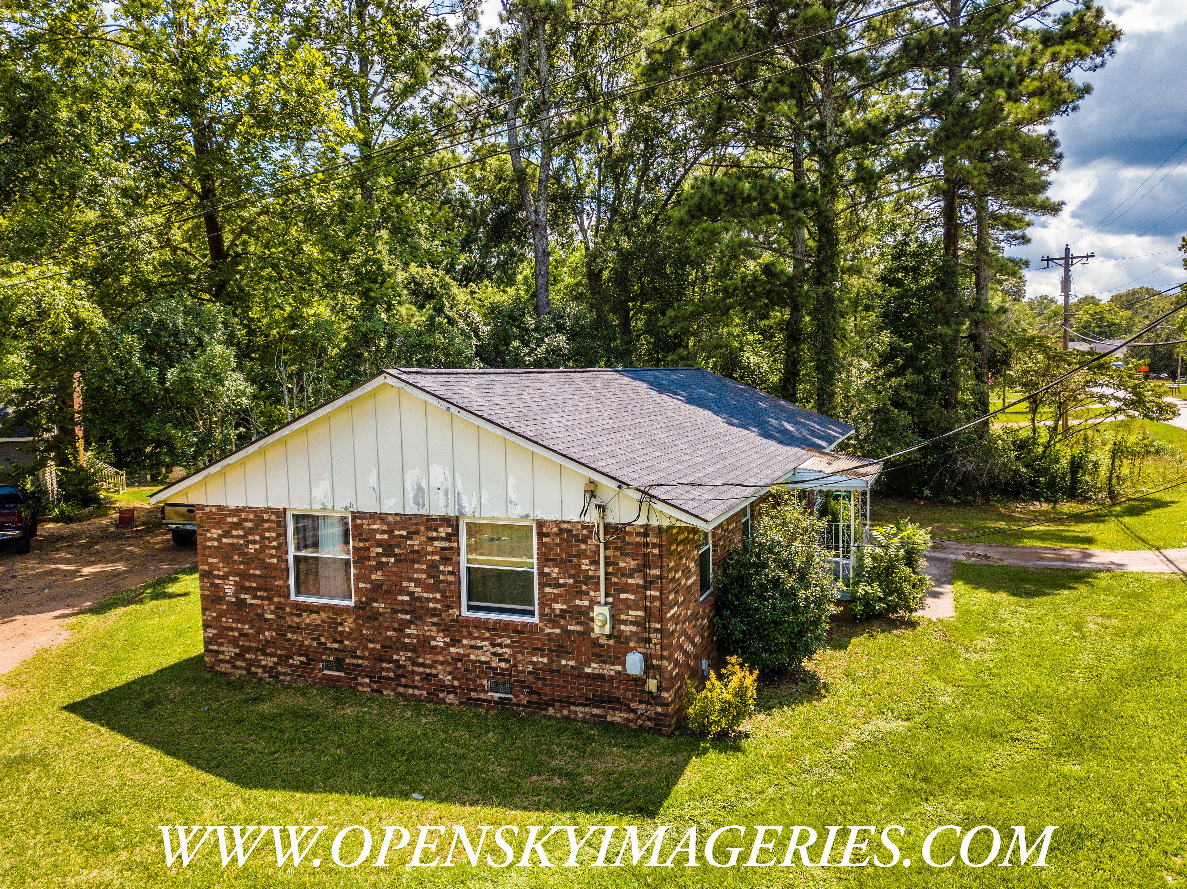 Oblique aerial drone view of a single-story brick residence with a gray shingle roof, white gable siding, green lawn, driveway, and mature trees surrounding the property.