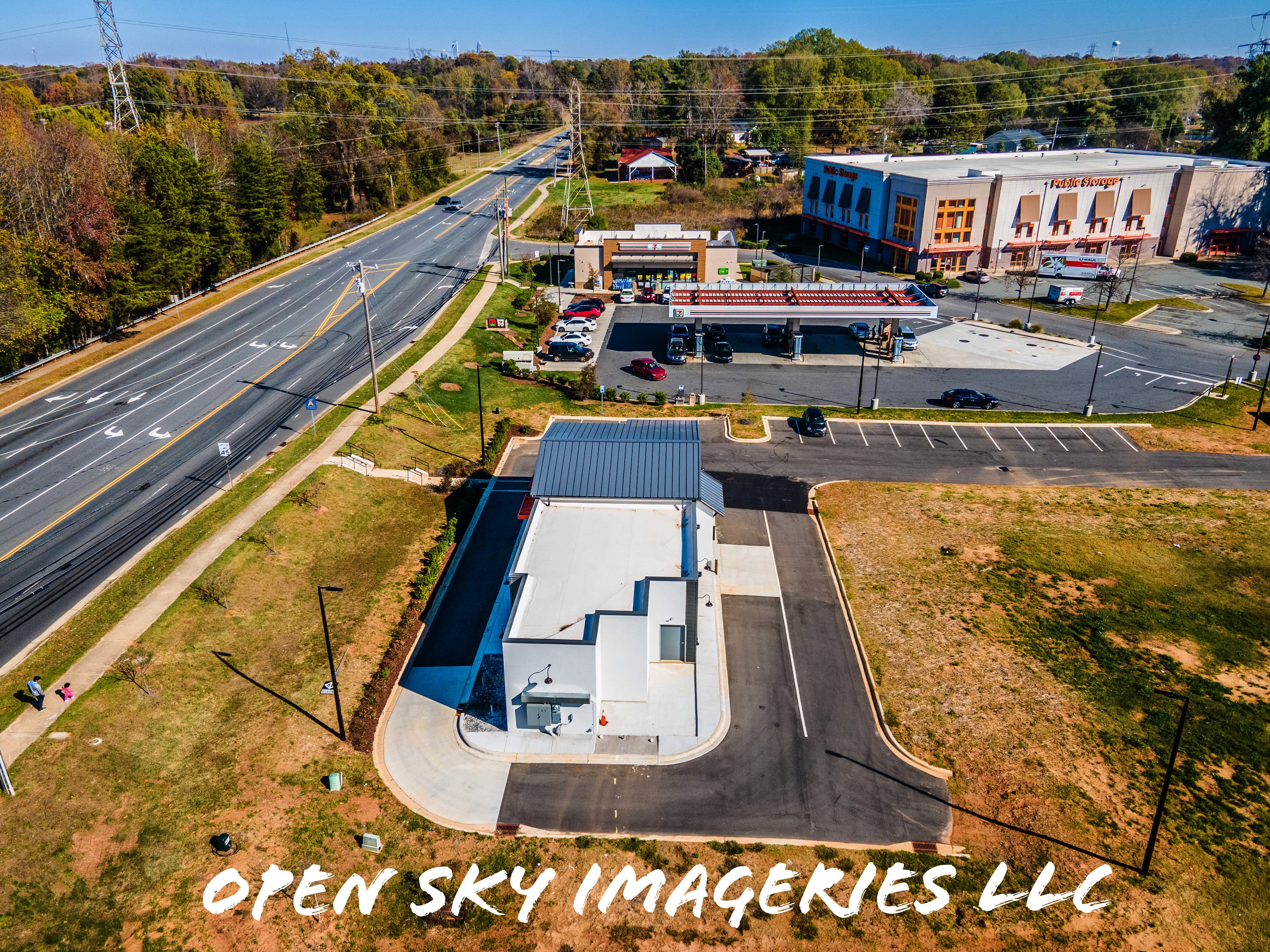 Aerial drone photograph of a commercial development featuring a standalone building with drive-through lanes, a 7-Eleven convenience store and fuel station, a Public Storage facility, parking areas, and a nearby roadway.