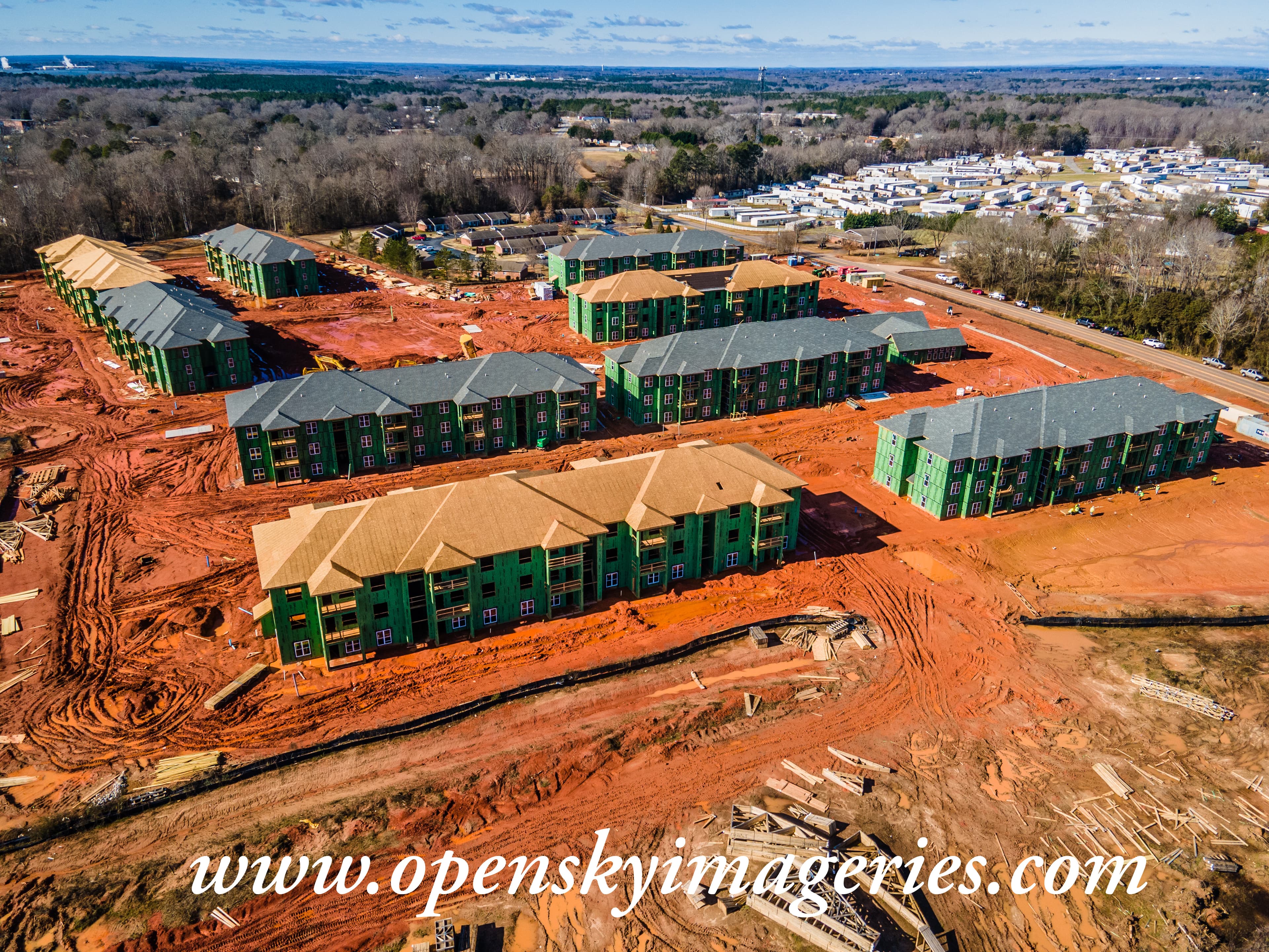 Aerial drone photograph of a large multifamily apartment development under construction, showing multiple three-story residential buildings with green exterior sheathing, partially completed roofs, red clay grading, construction materials, and active site preparation.