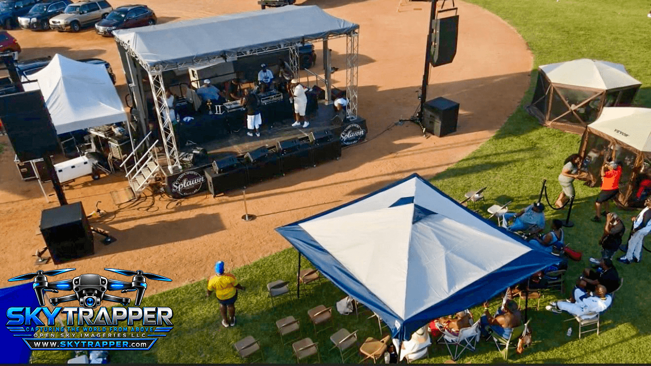 Elevated aerial view of an outdoor DJ event with performers on a covered stage, sound equipment, event tents, and attendees seated beneath a canopy.
