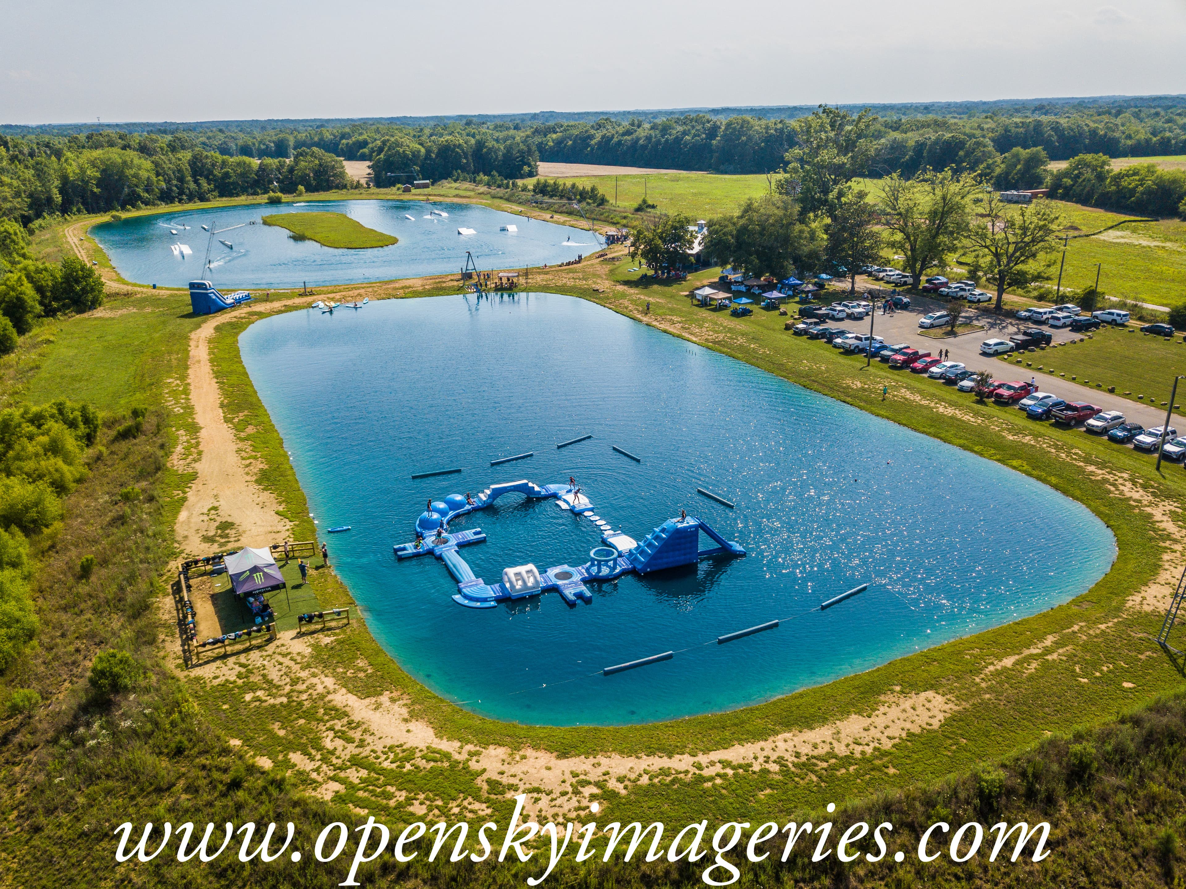 Aerial drone photograph of an outdoor water recreation park featuring two large ponds, floating inflatable obstacle courses, water slides, visitors, event tents, and a parking area surrounded by green fields and woodland.