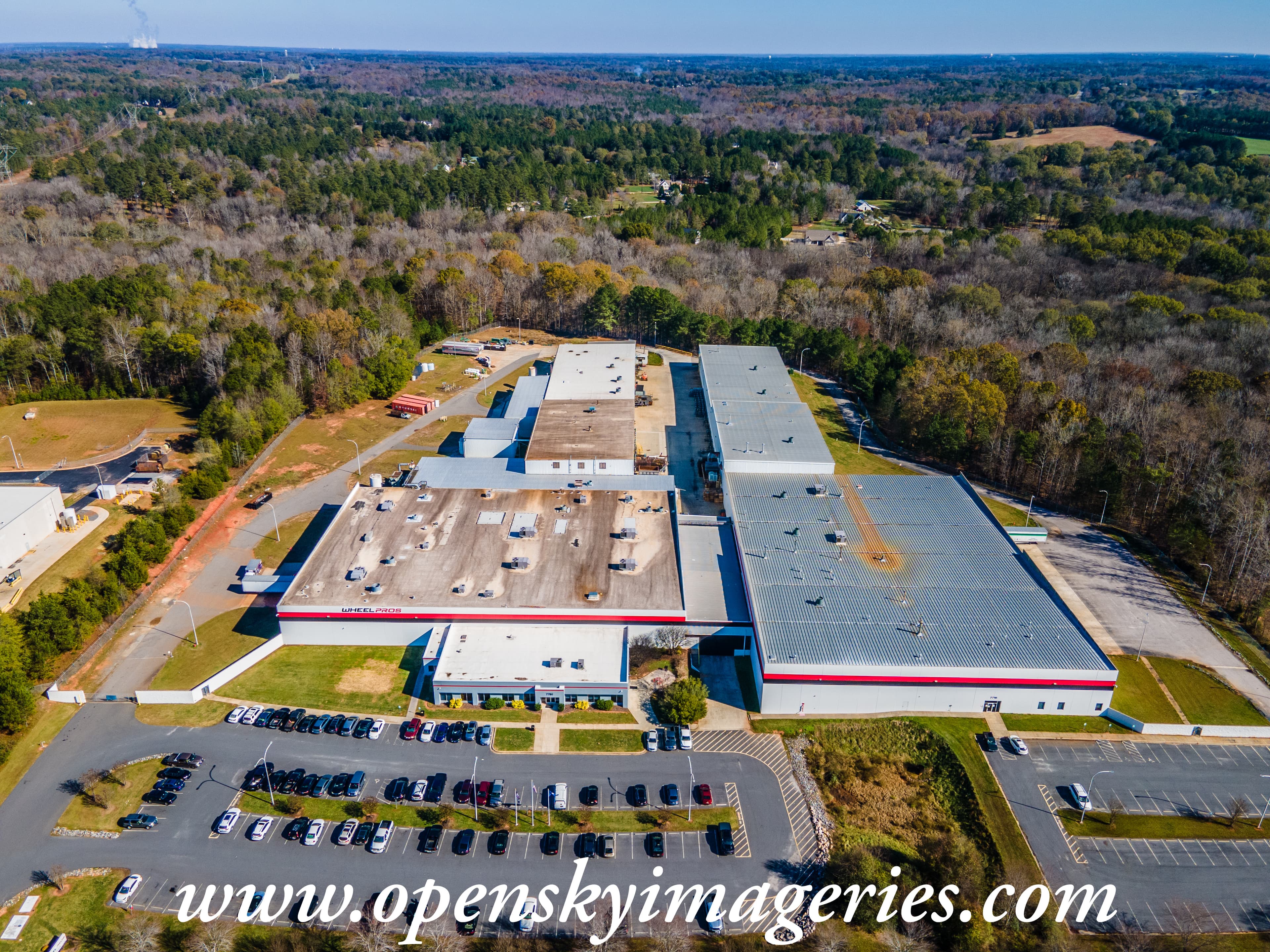 Aerial drone photograph of the Wheel Pros industrial facility featuring multiple large warehouse buildings, expansive rooftops, employee parking areas, access roads, and surrounding wooded landscape.