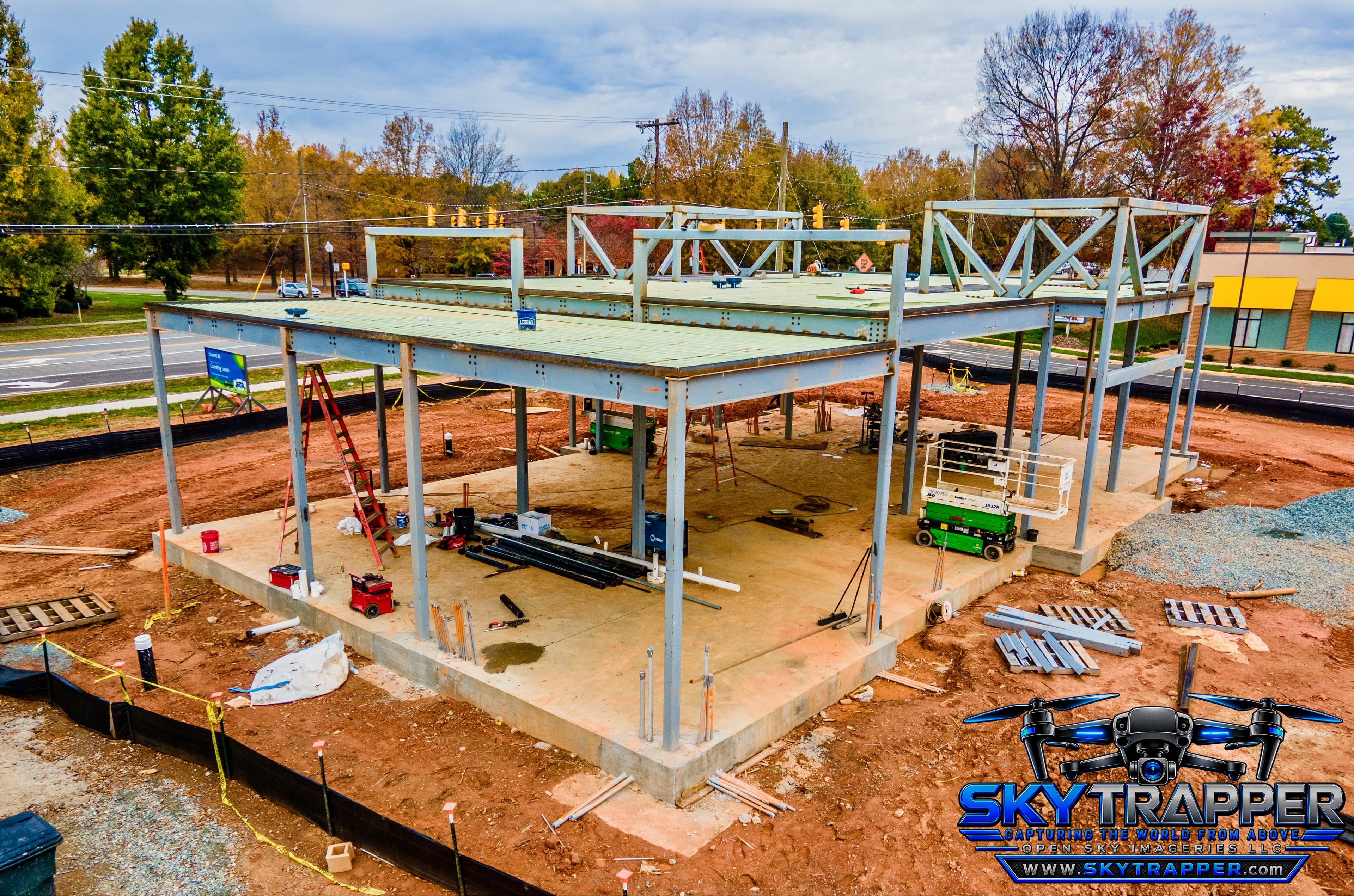 Aerial drone photograph of a commercial building under construction, showing structural steel framing, elevated metal decking, concrete foundations, construction equipment, and building materials at an active jobsite.