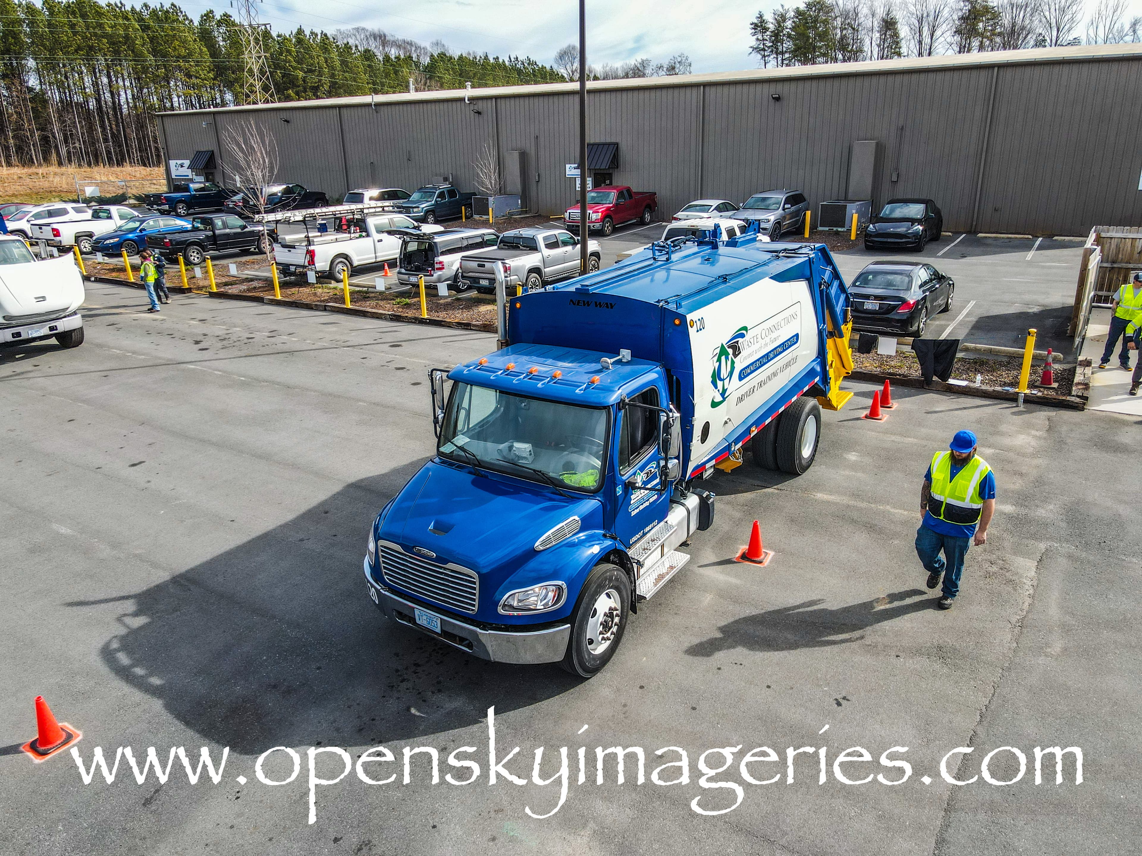 Elevated drone photograph of a blue commercial waste collection truck parked outside an industrial facility, with employees in safety vests, parked vehicles, and a warehouse building.