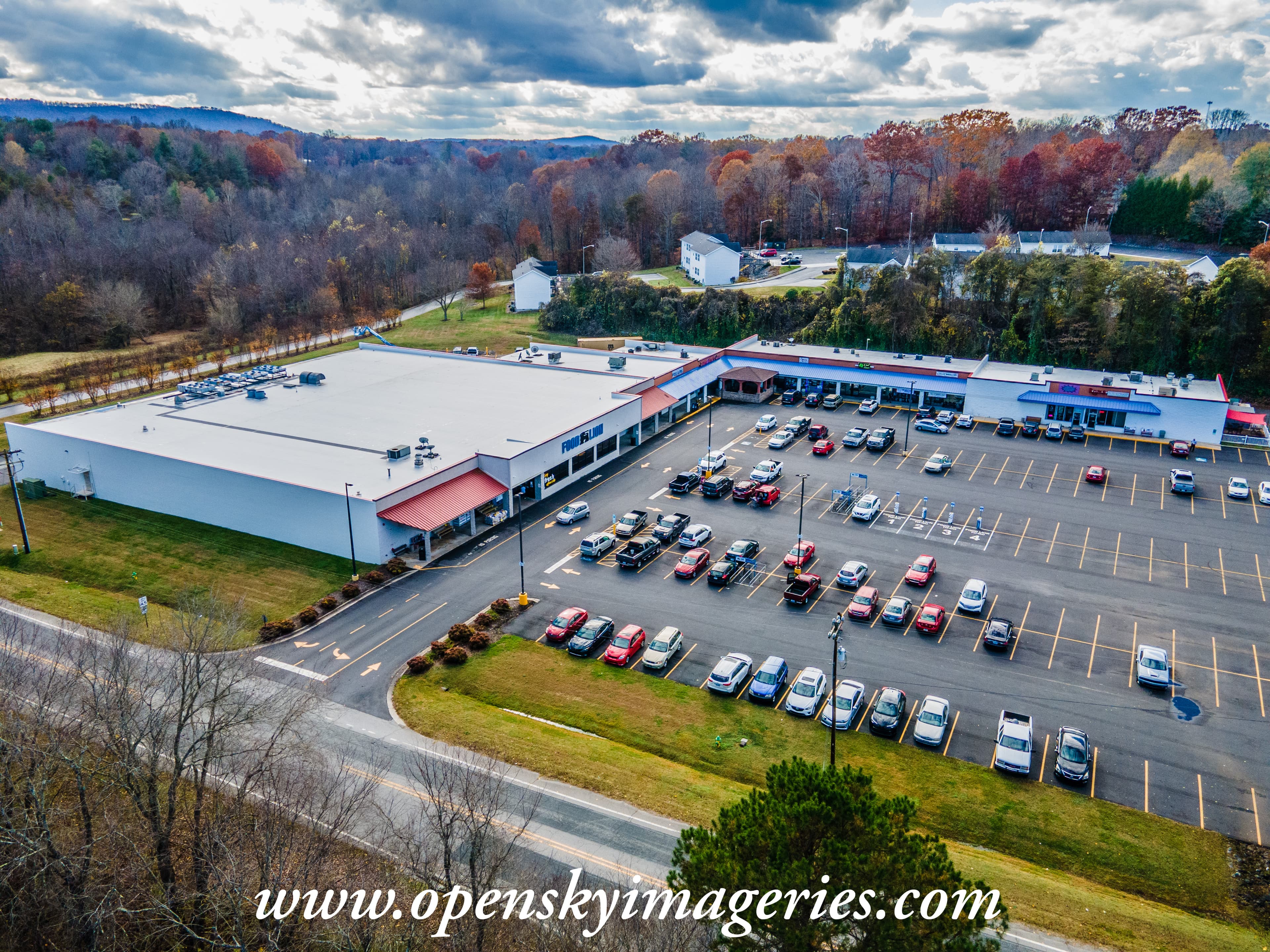Aerial drone photograph of a suburban shopping center featuring a Food Lion grocery store, adjoining retail storefronts, a large parking lot, surrounding residential properties, and wooded hills with autumn foliage.