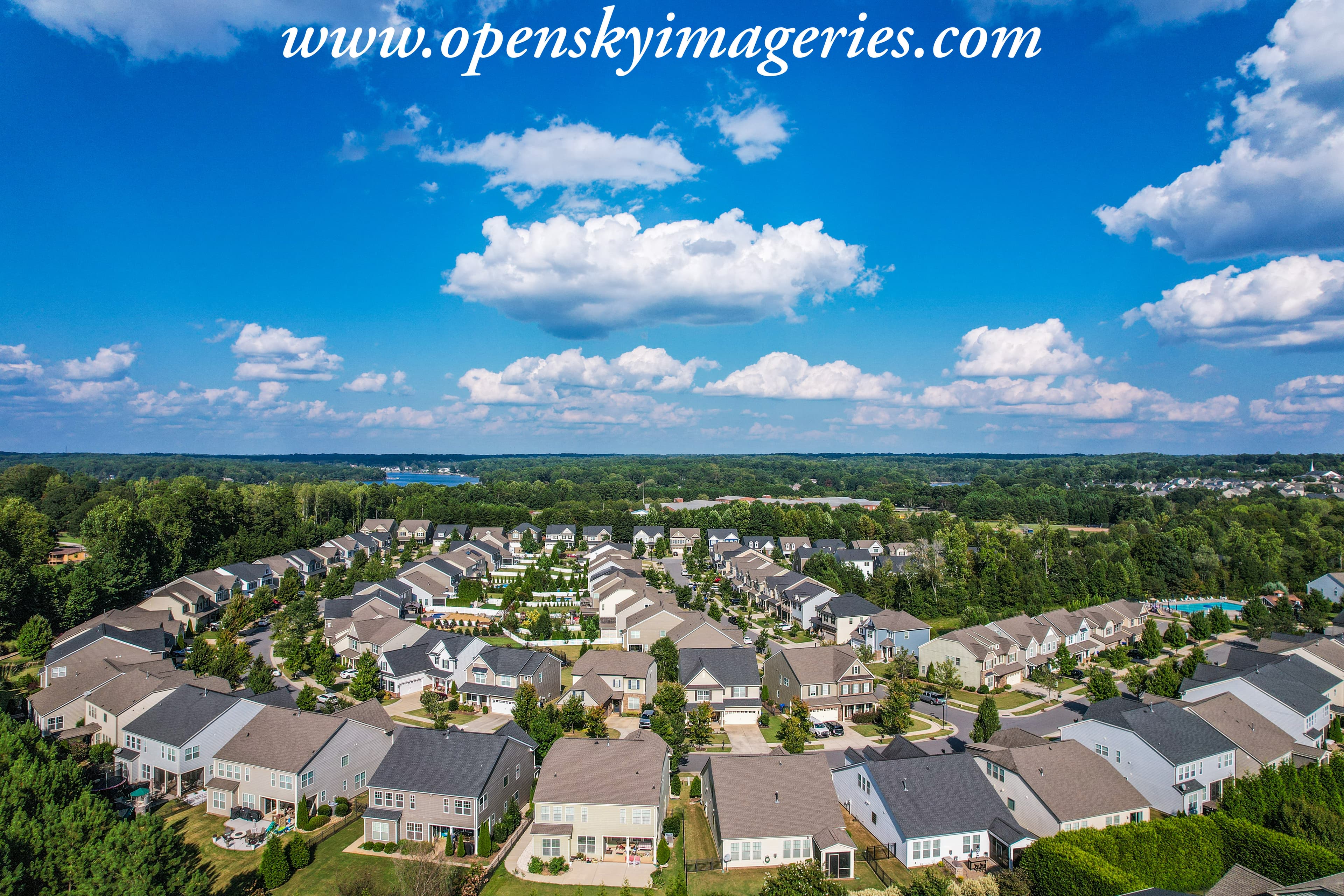 Wide aerial drone photograph of a suburban residential neighborhood featuring rows of detached houses, landscaped yards, tree-lined streets, a community swimming pool, and surrounding woodland beneath a bright blue sky with scattered white clouds.