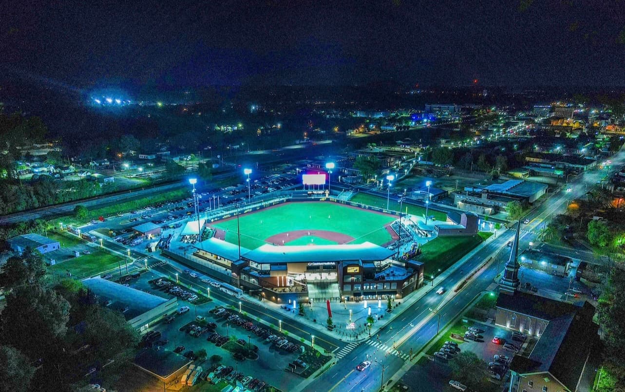 Nighttime aerial drone photograph of an illuminated baseball stadium, showing the brightly lit playing field, grandstands, stadium buildings, surrounding streets, parking areas, and city lights.