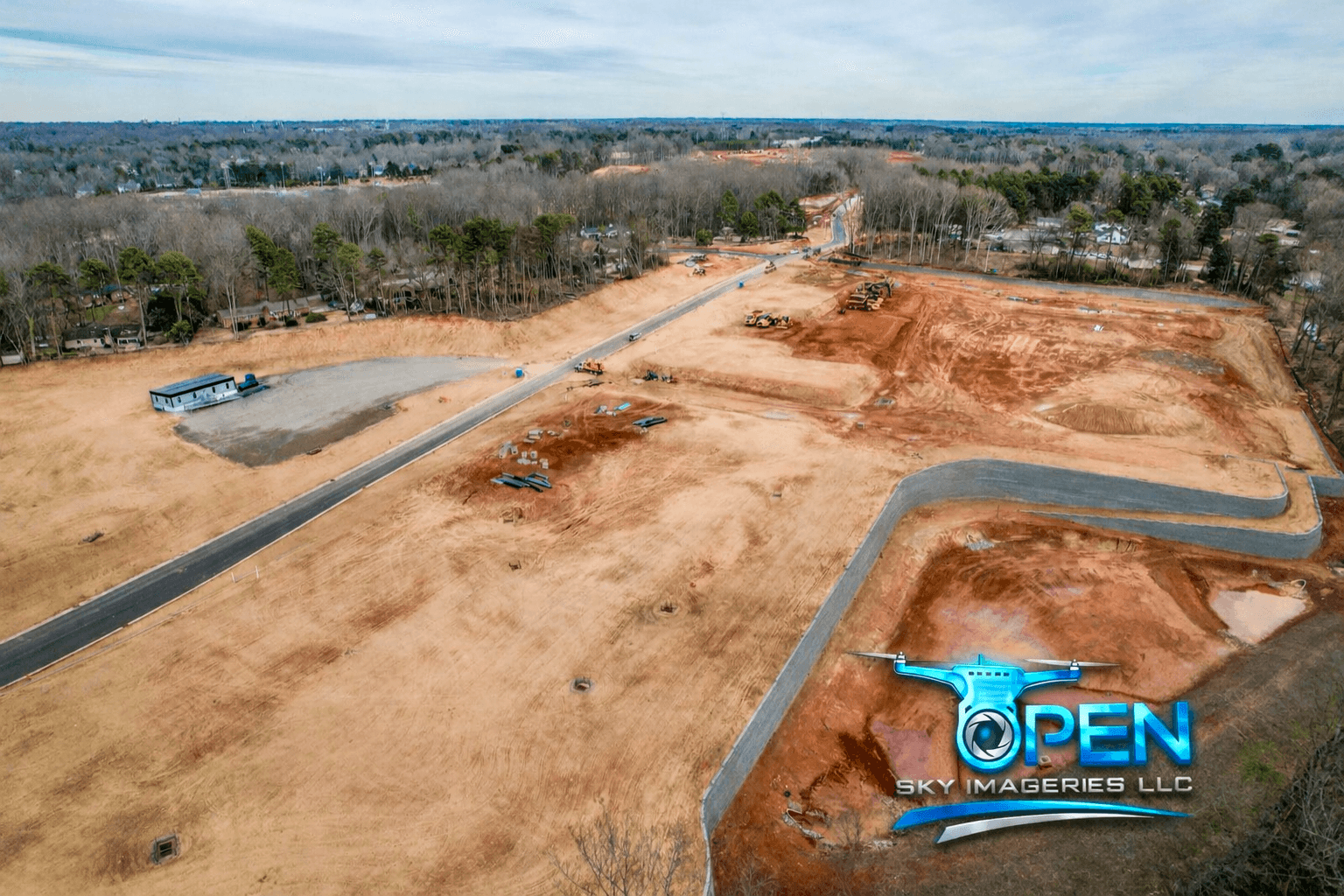 Wide aerial view of a large land development site showing graded red-clay terrain, newly paved roads, earthmoving equipment, a temporary site trailer, and surrounding wooded residential areas.