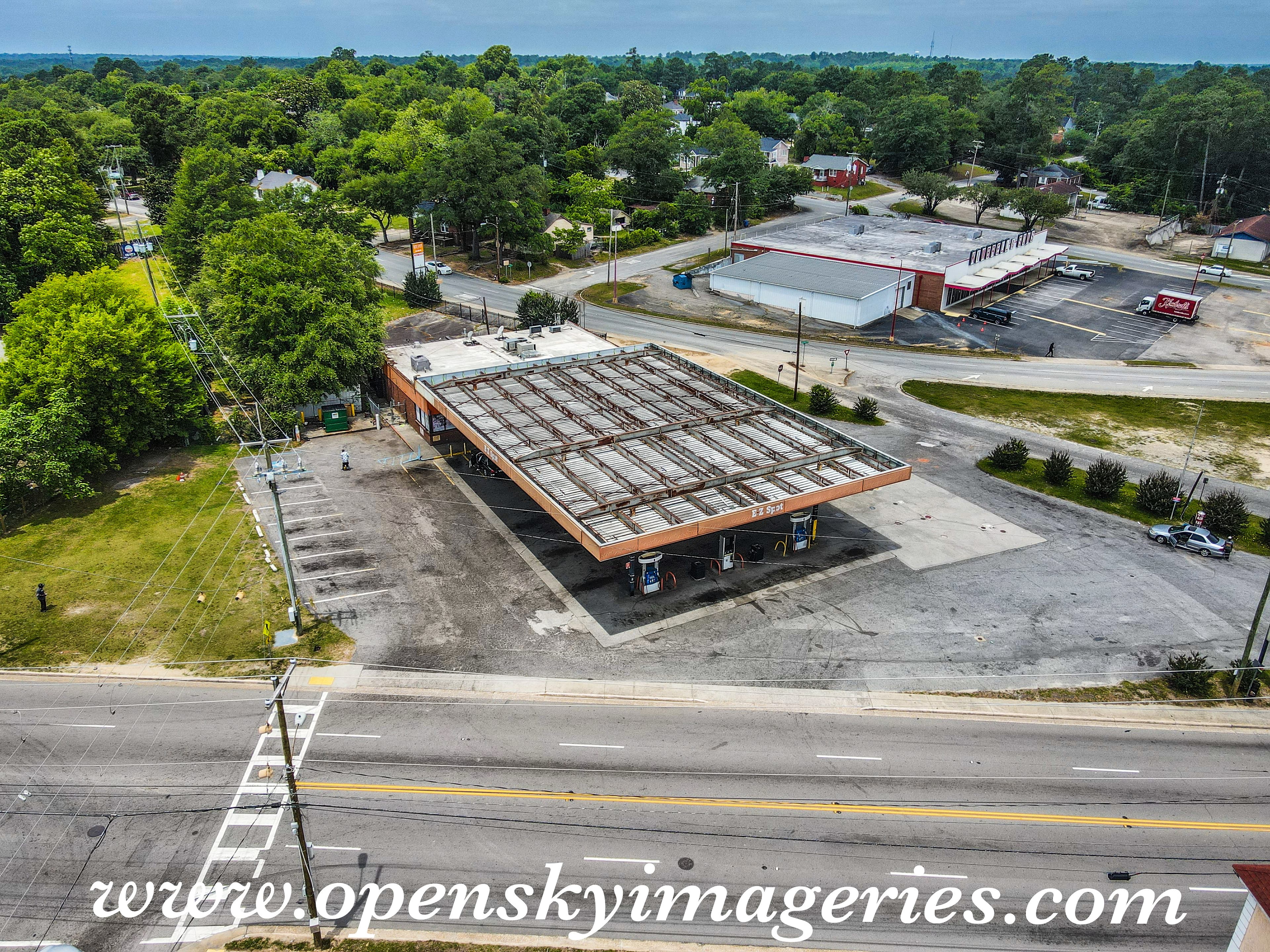 Aerial drone photograph of a roadside gas station property featuring a large metal canopy over fuel pumps, an attached brick building, paved parking areas, and surrounding commercial and residential properties.