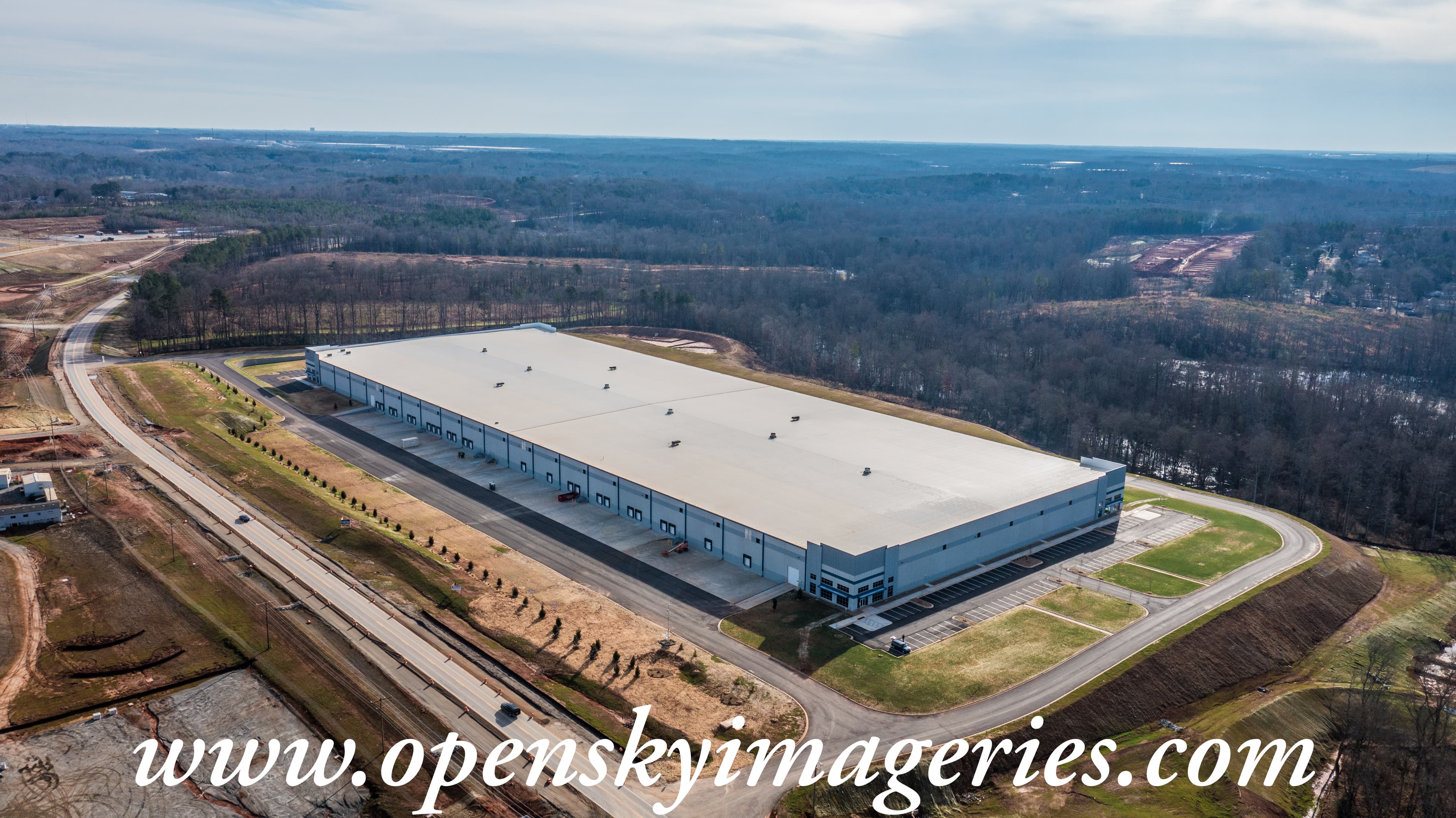 Aerial drone photograph of a large industrial warehouse and distribution facility featuring multiple loading dock doors, an expansive flat roof, paved access roads, landscaped grounds, and surrounding wooded property.