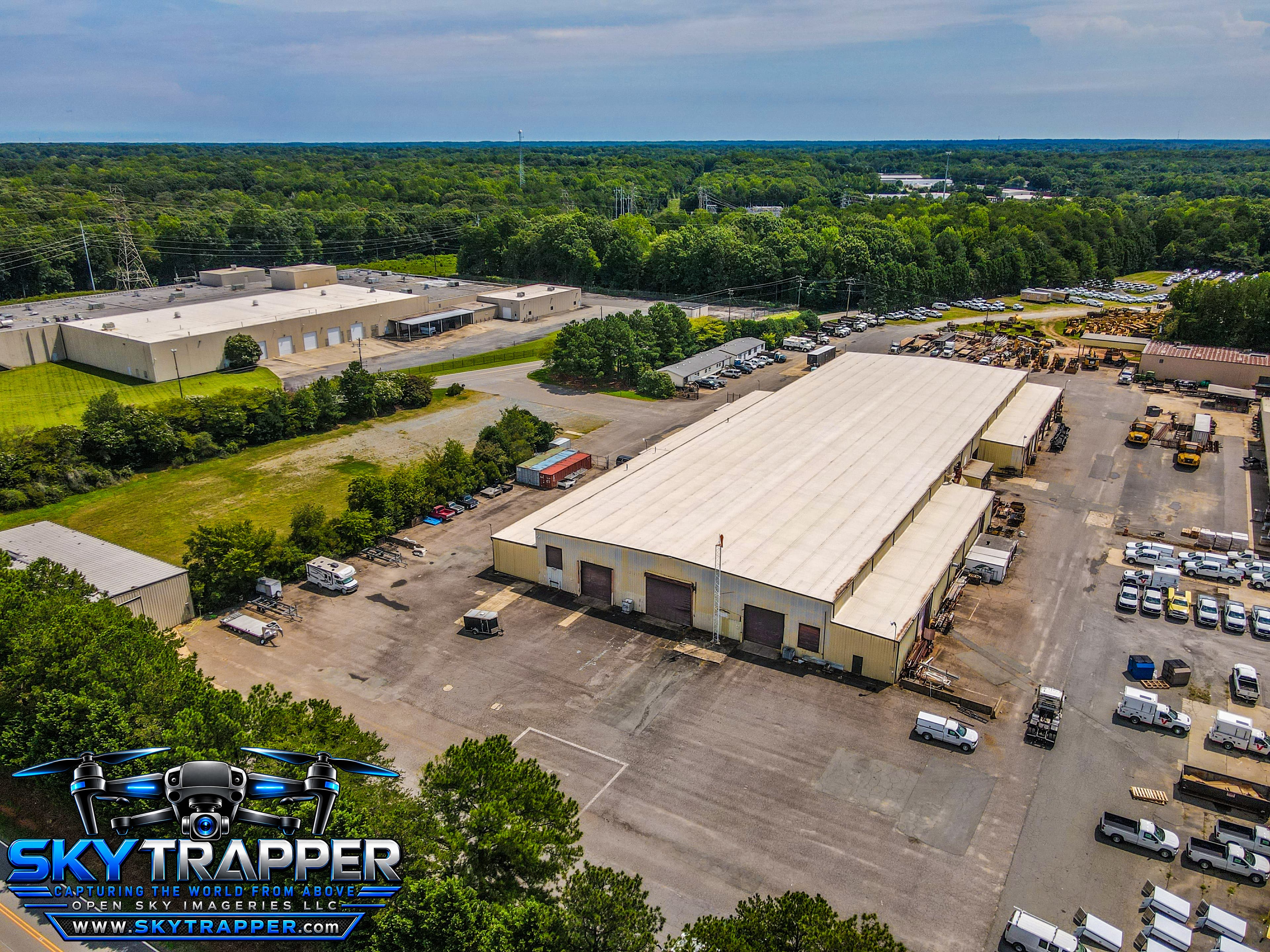Aerial drone photograph of a large industrial warehouse and equipment yard featuring multiple loading bays, service trucks, heavy machinery, adjacent commercial buildings, and surrounding wooded areas.