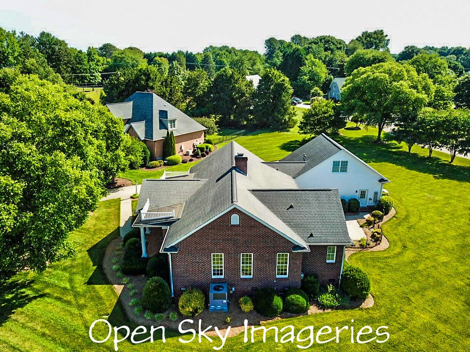 Elevated drone view of a brick suburban residence with gray shingle roof, attached white exterior section, landscaped gardens, green lawn, and mature trees.