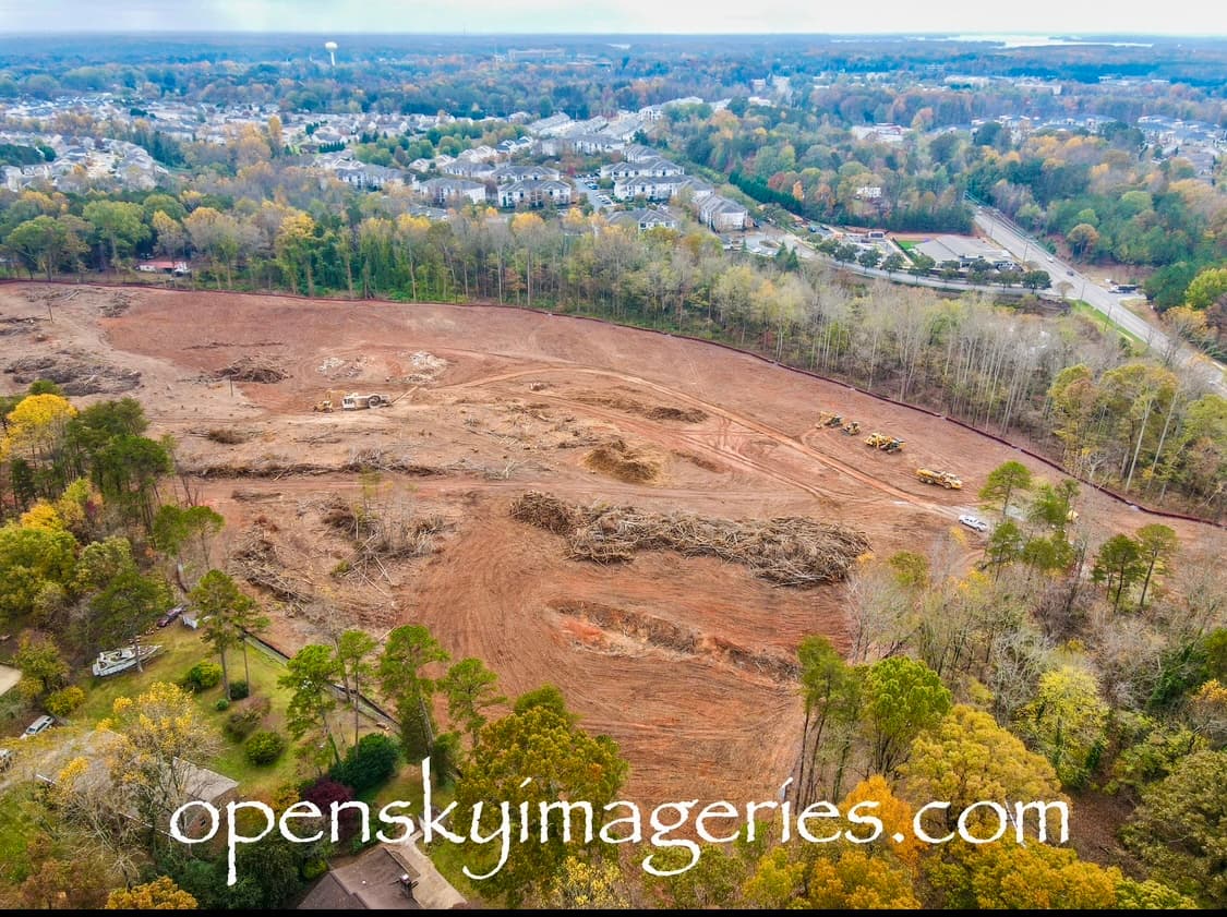 Aerial view of an active land-clearing and grading site with earthmoving equipment, cleared vegetation, and nearby residential neighborhoods.