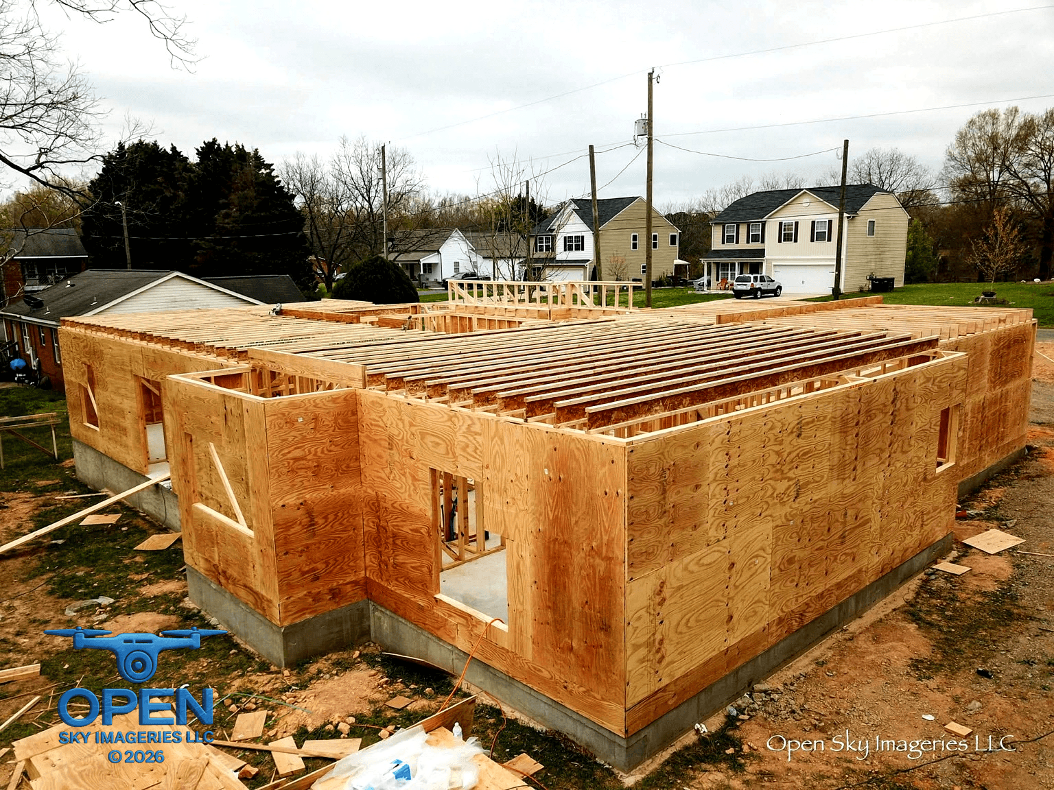 Elevated drone photograph of a residential building under construction, showing plywood-sheathed exterior walls, exposed wooden joists, window openings, and a concrete foundation in an established neighborhood.