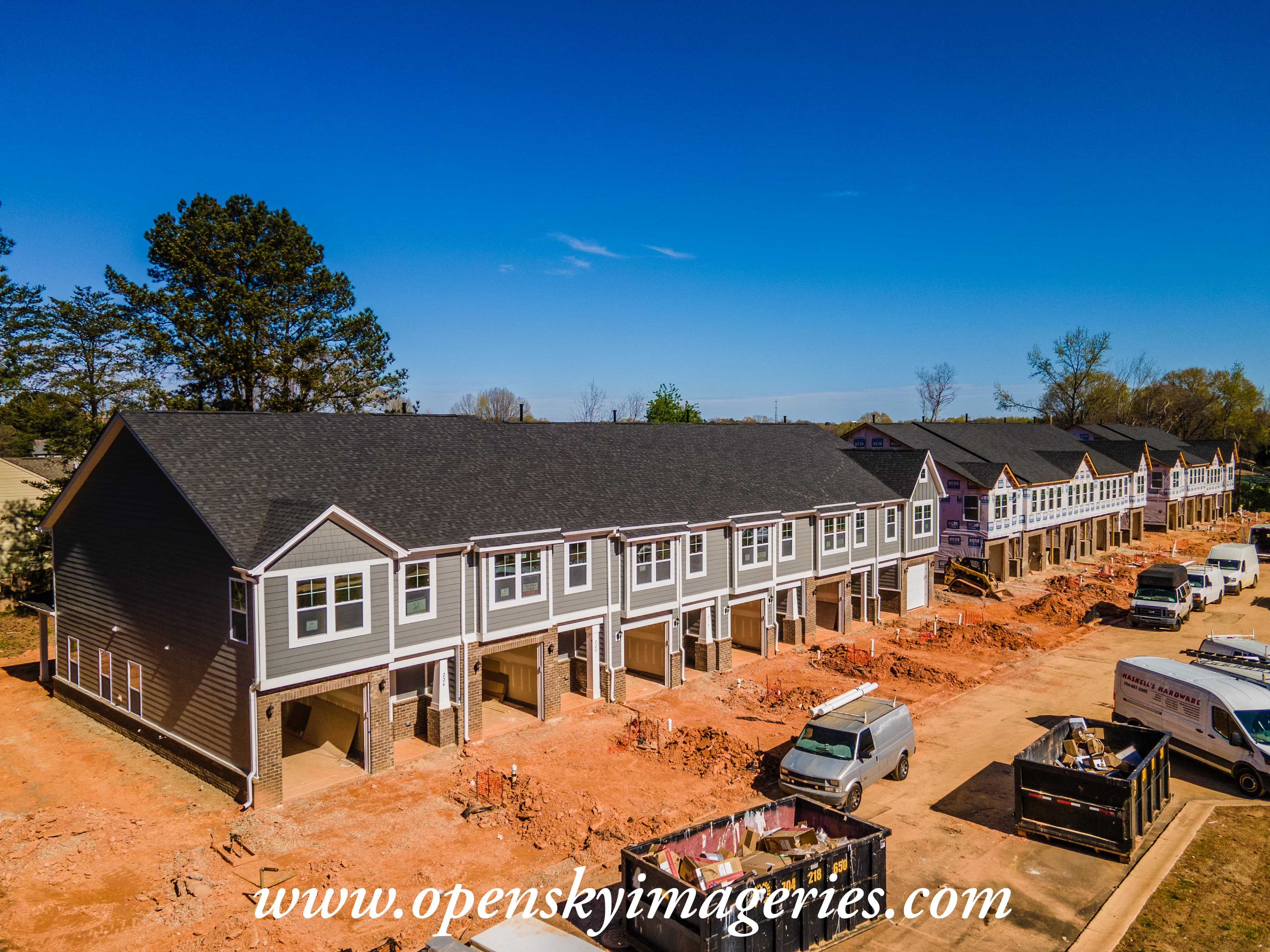 Aerial drone photograph of a residential townhome development under construction, showing a row of two-story attached homes with installed roofs, partially completed exteriors, open garages, construction vehicles, and red clay grading.