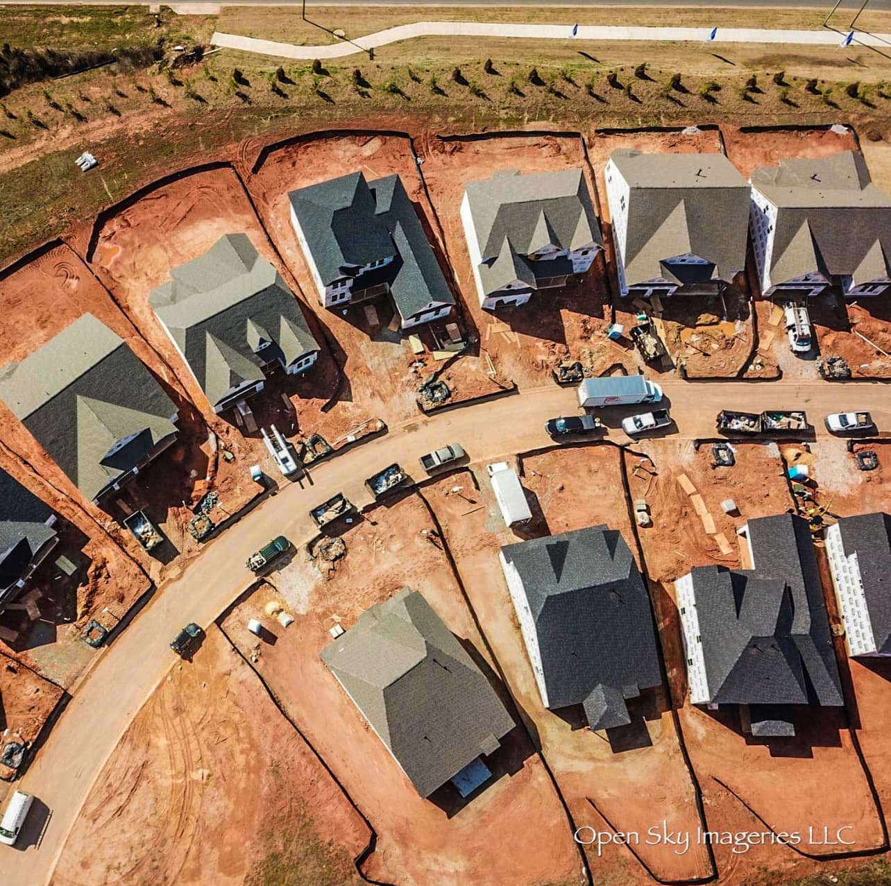 Overhead drone photograph of a residential subdivision under construction, showing newly built houses at different completion stages, shingle roofs, red clay lots, construction equipment, and vehicles along a curved neighborhood road.