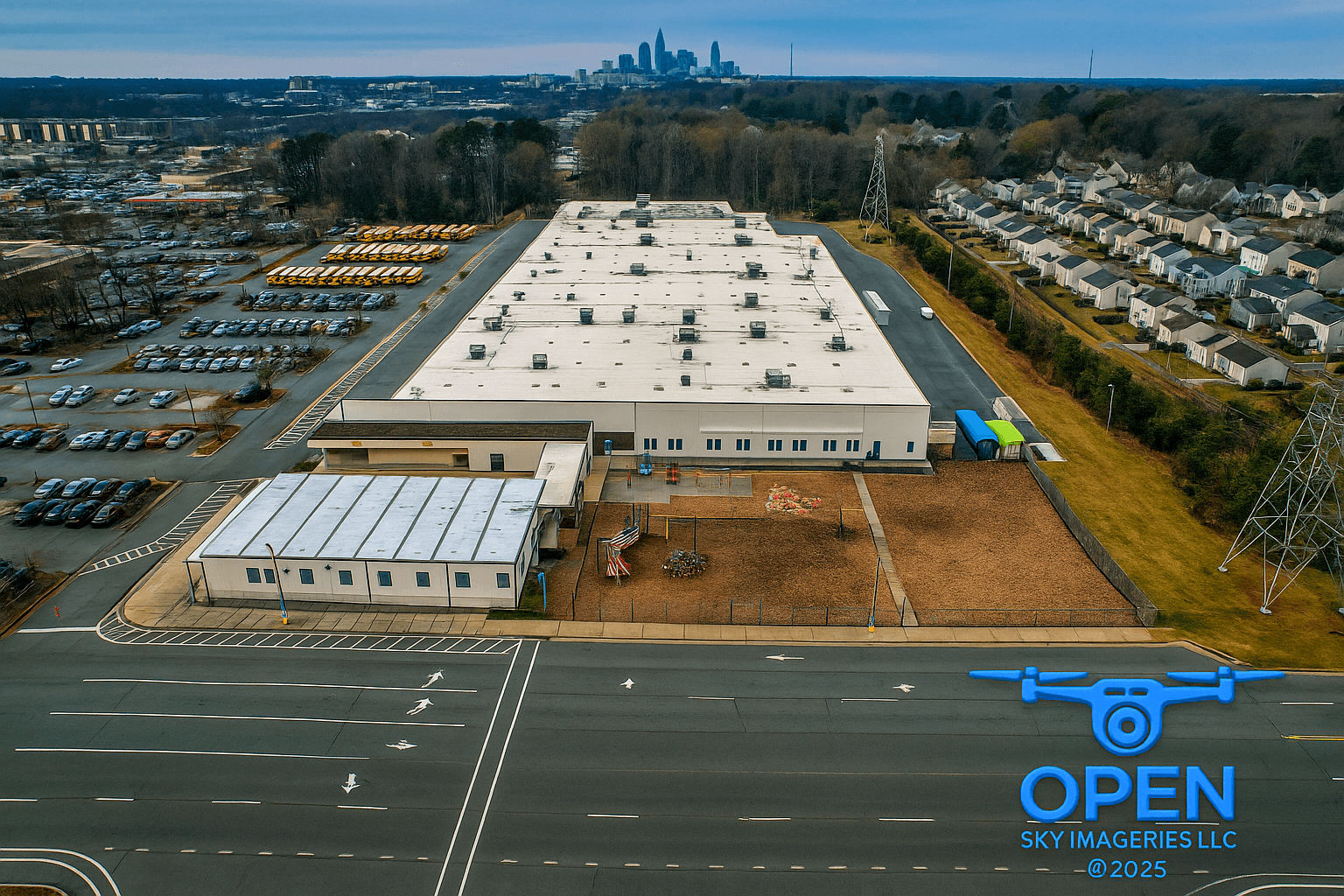 Aerial view of a large industrial facility with a flat white roof, extensive vehicle parking and storage areas, adjacent residential neighborhoods, and the Charlotte skyline in the distance.