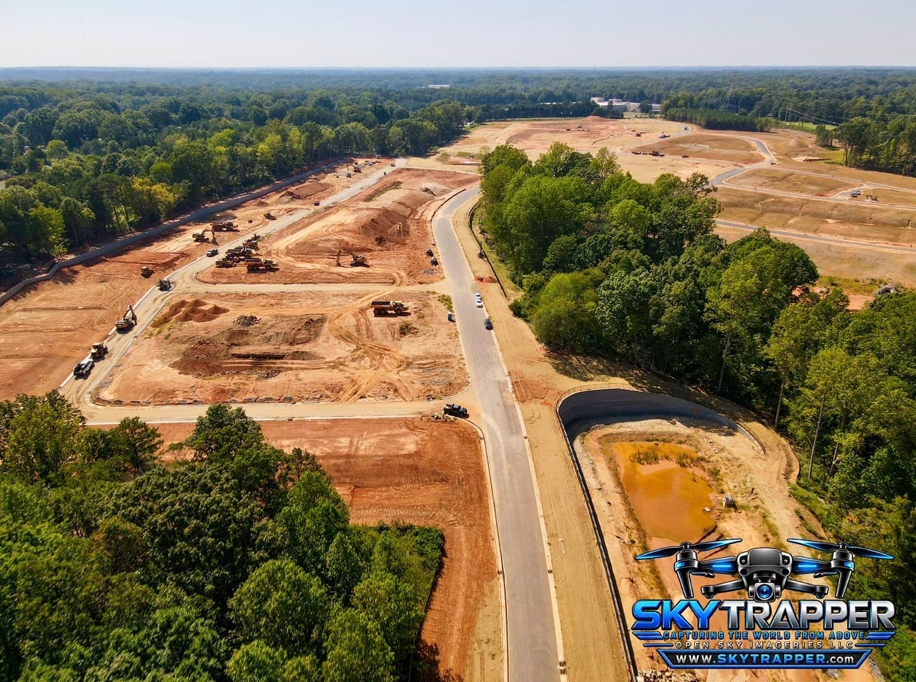 Aerial drone photograph of a large construction development featuring earthmoving equipment, graded building sites, newly paved roads, stormwater infrastructure, and surrounding woodland.