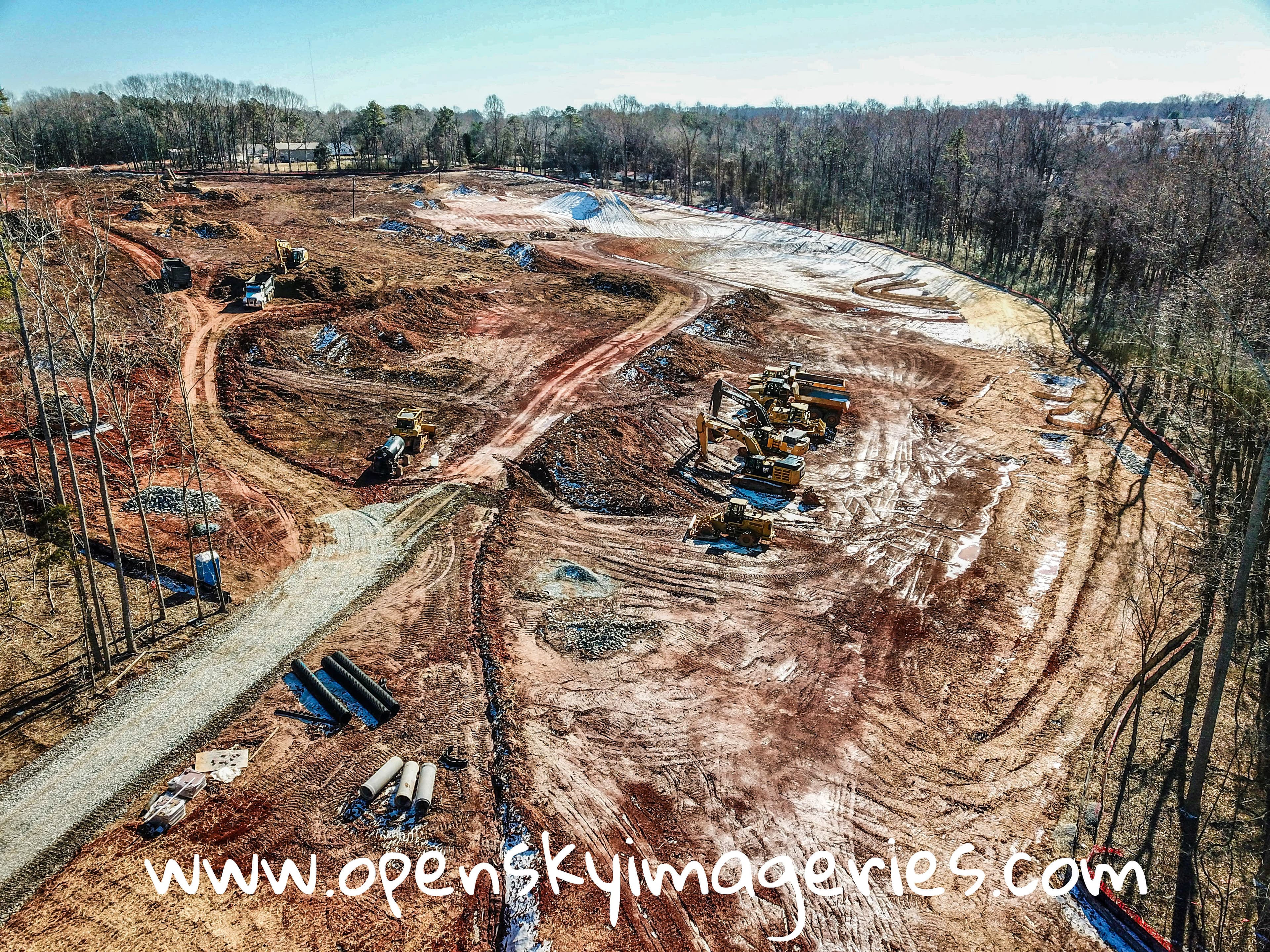Drone aerial view of an active construction site with excavators, bulldozers, dump trucks, graded earth, access roads, and drainage pipes surrounded by woodland.