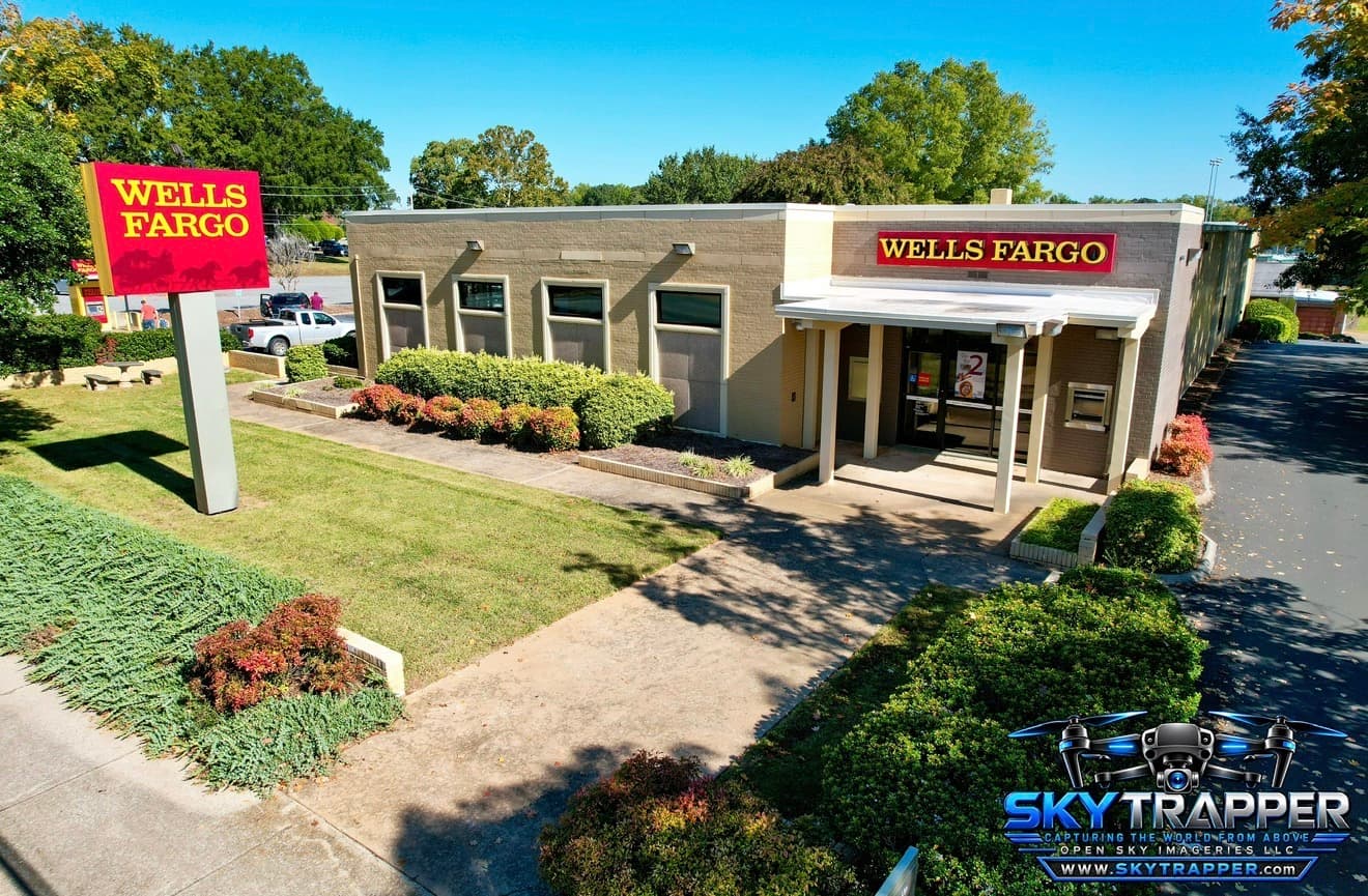 Elevated aerial photograph of a Wells Fargo bank branch with prominent red-and-yellow signage, covered front entrance, ATM, landscaped lawn, shrubs, and adjacent parking area.