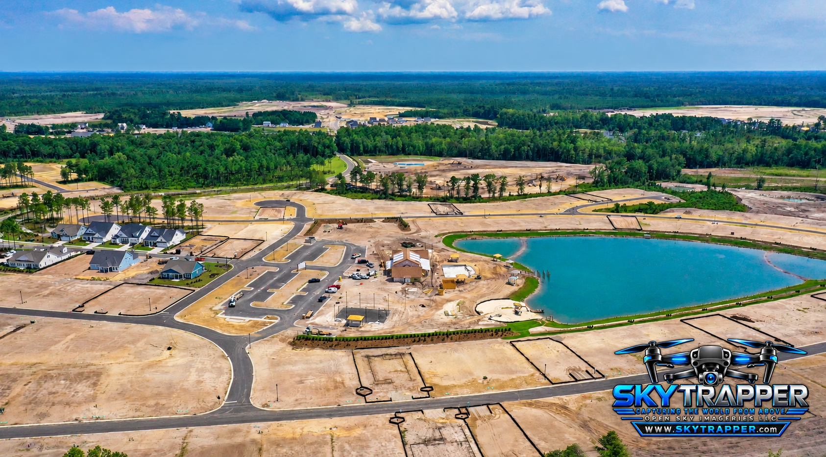 Wide aerial photograph of a residential development under construction, showing newly paved roads, graded homesites, completed houses, a large pond, and surrounding wooded areas.