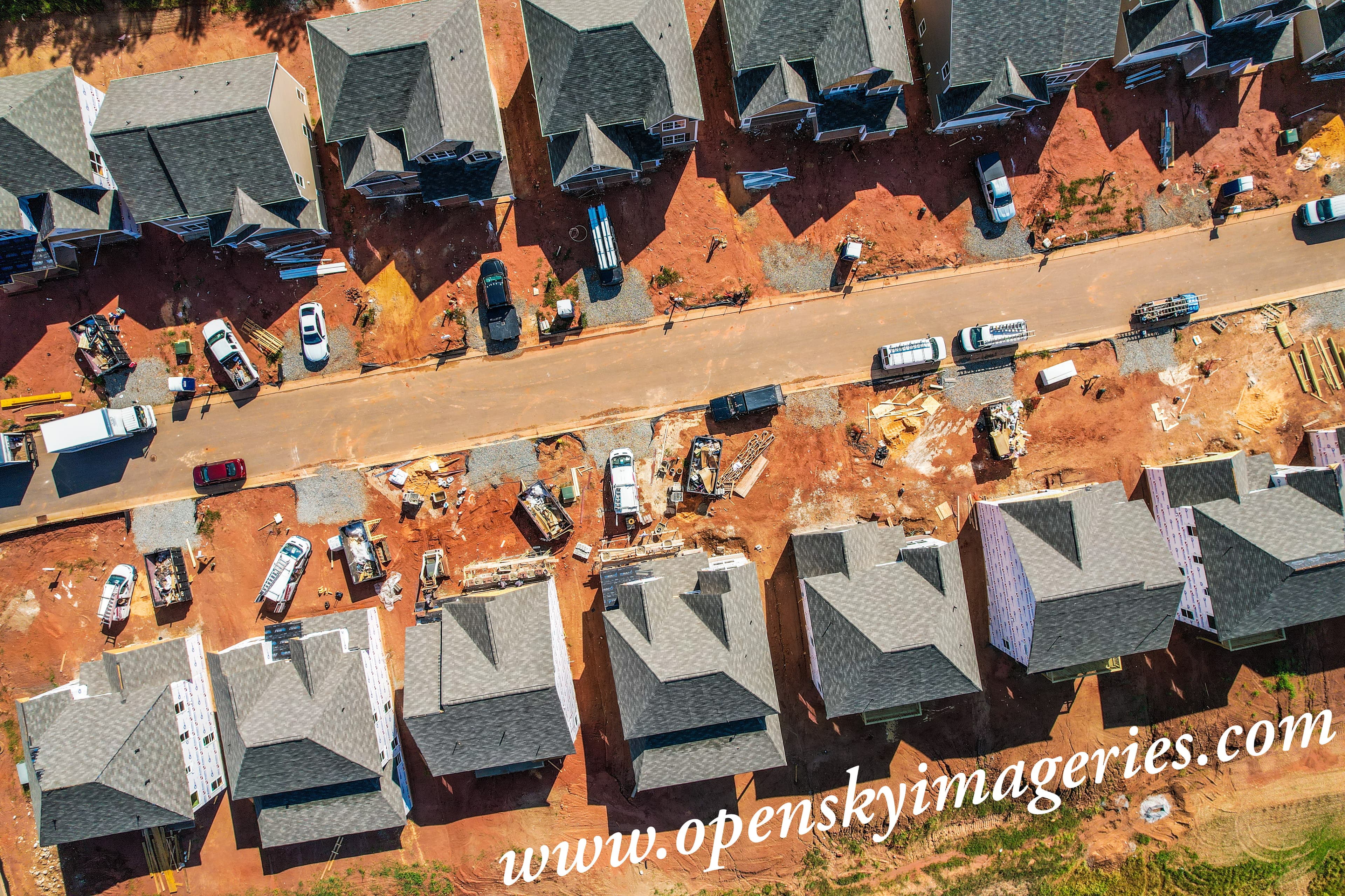 Top-down aerial drone photograph of a residential construction development showing rows of newly built houses, shingled roofs, partially finished exteriors, construction equipment, work vehicles, building materials, and a paved access road.
