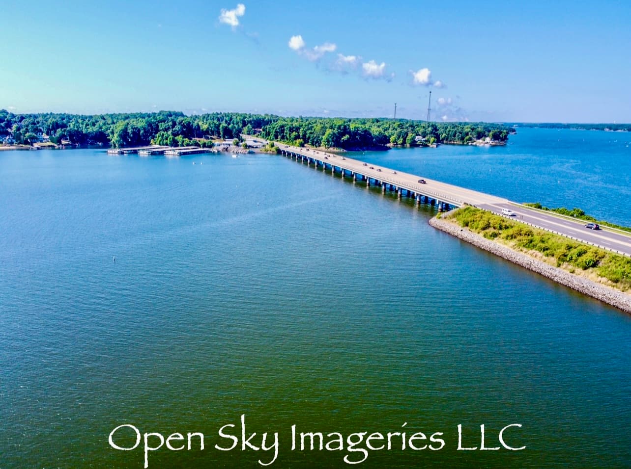 Aerial drone photograph of a long highway bridge crossing a wide blue lake, with vehicles traveling across the bridge, wooded shorelines, waterfront homes, and clear summer skies.