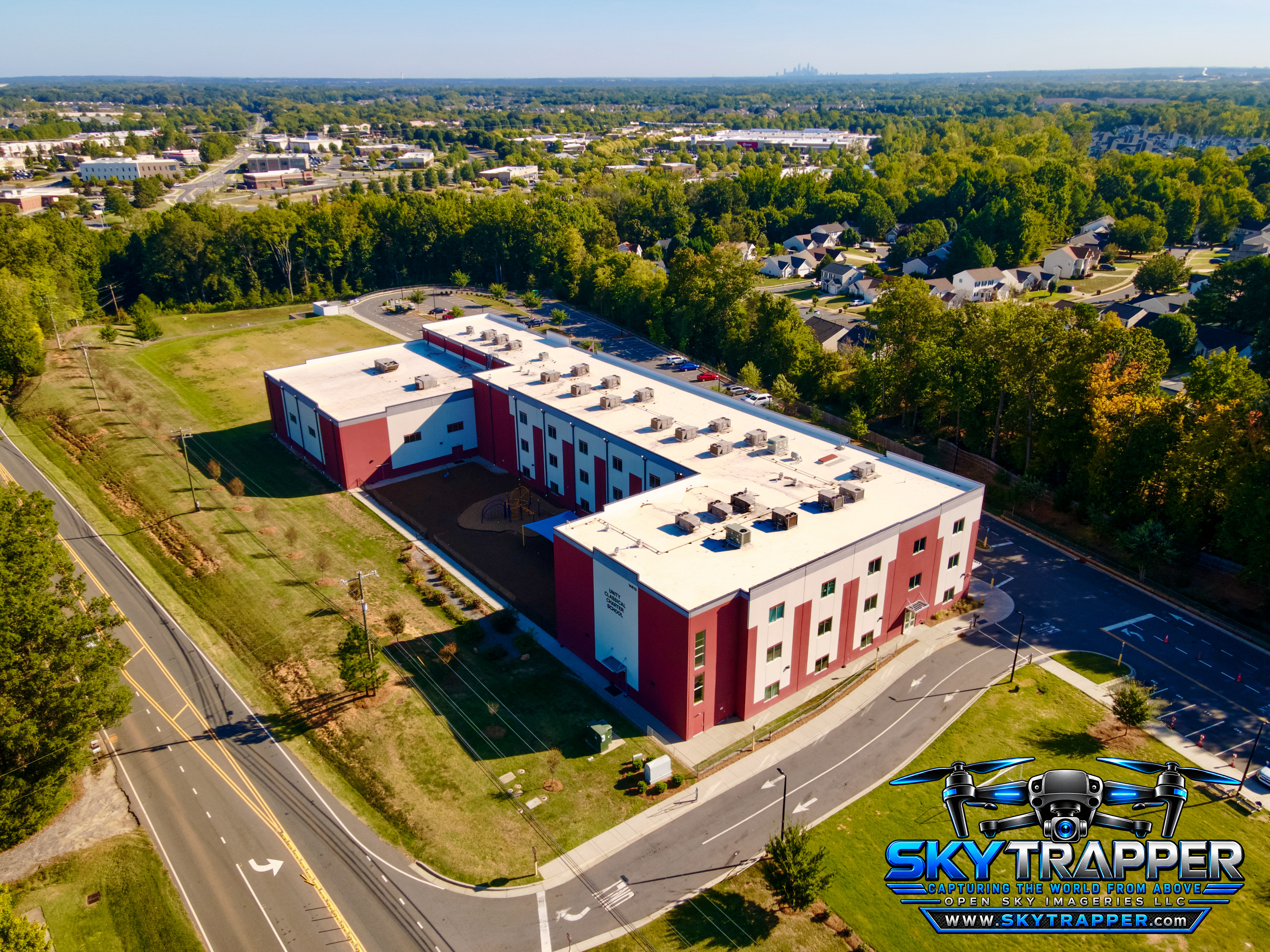Oblique drone photograph of a large red-and-white multistory building with flat rooftops, landscaped grounds, parking areas, surrounding roads, and nearby residential neighborhoods.