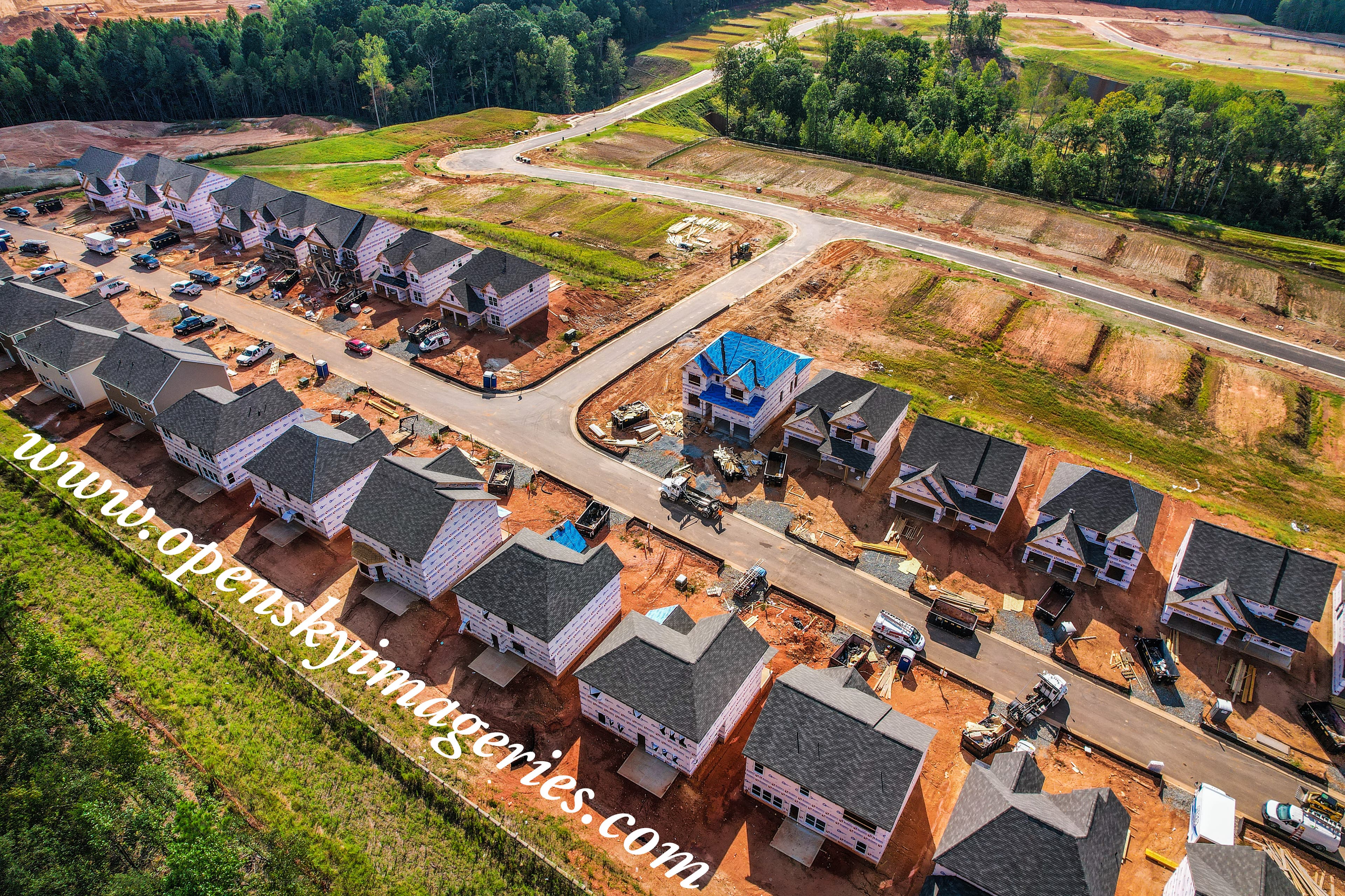 Oblique aerial drone photograph of a residential subdivision under construction, showing rows of houses at various building stages, paved roads, construction vehicles, building materials, graded lots, and surrounding woodland.