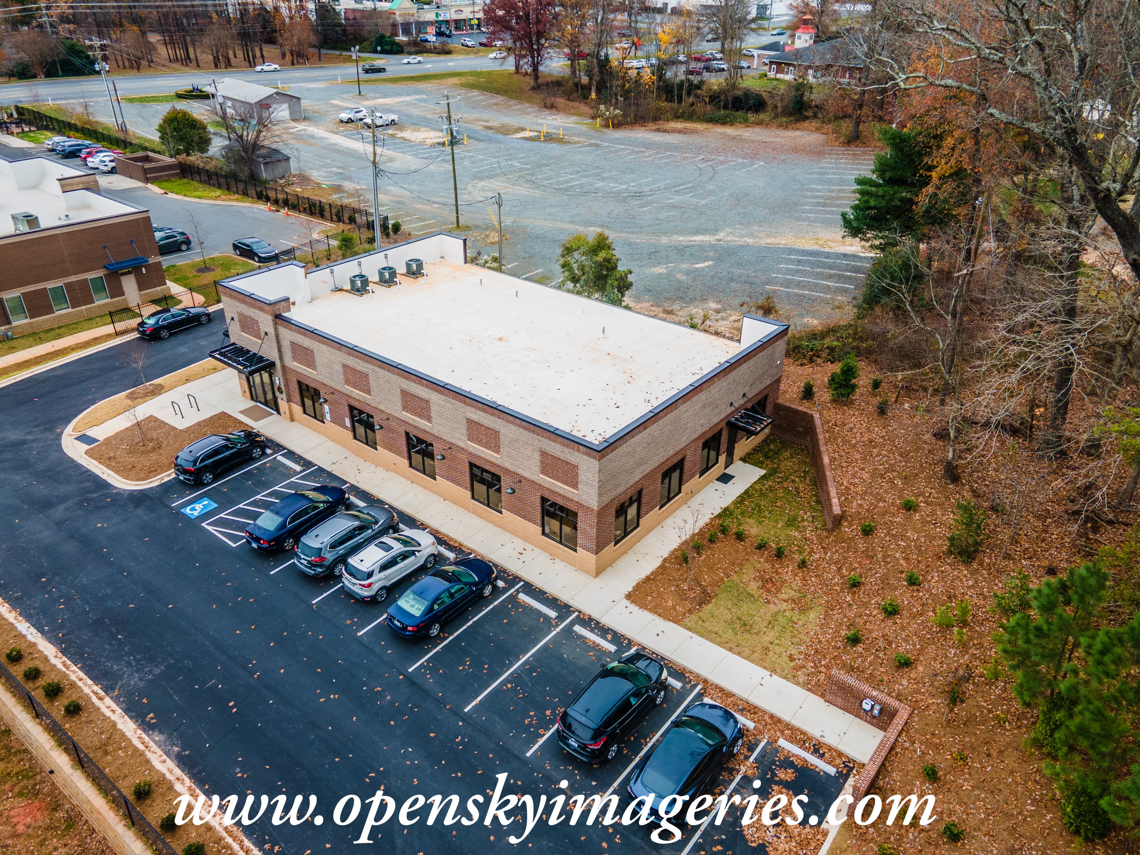 Aerial drone photograph of a single-story brick commercial building with a flat roof, paved parking lot, accessible parking spaces, landscaped grounds, and surrounding commercial properties.