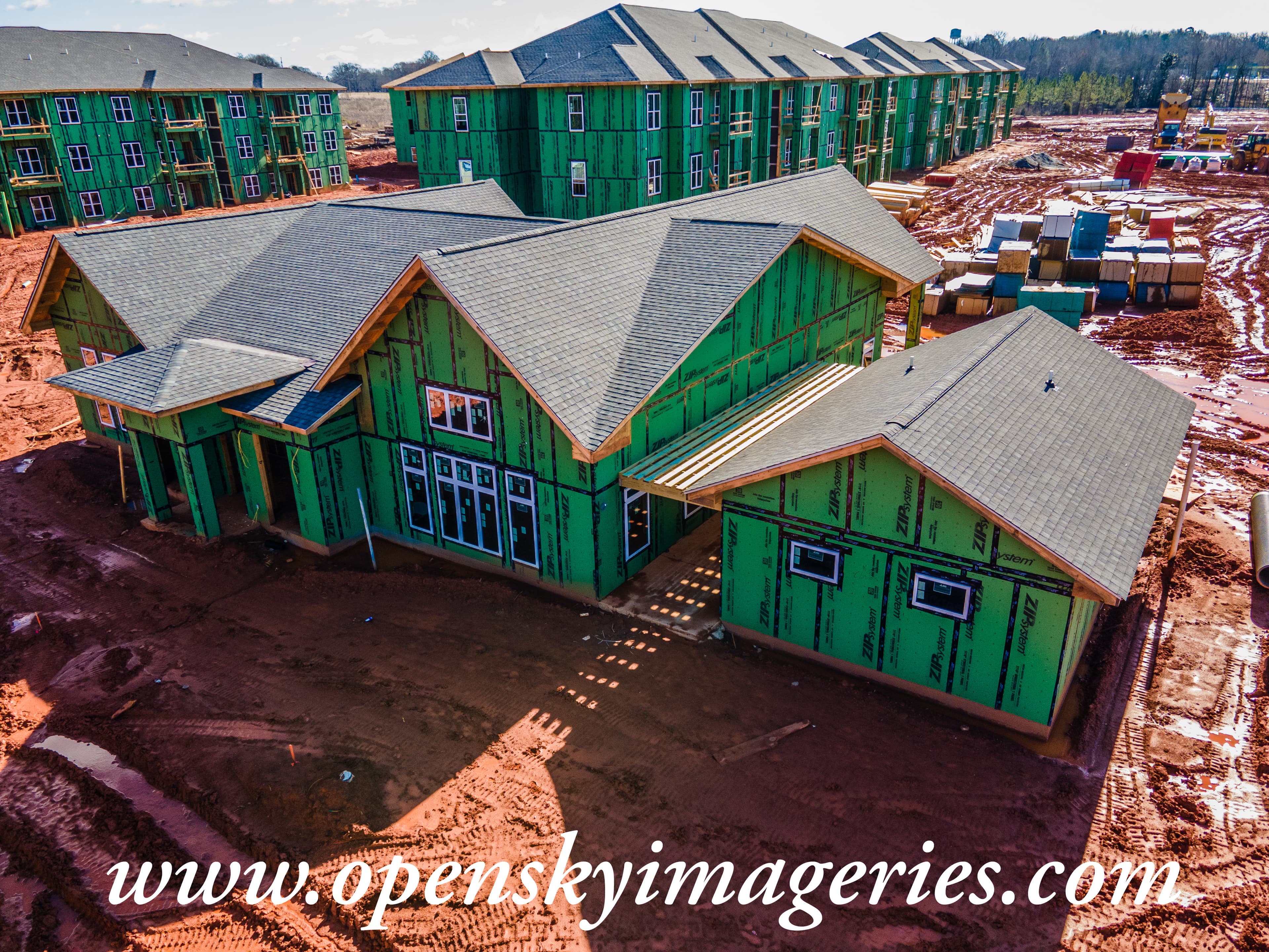 Aerial drone photograph of a multifamily residential construction site showing a large clubhouse-style building and three-story apartment buildings with green exterior sheathing, installed shingle roofs, unfinished balconies, construction materials, and red clay terrain.