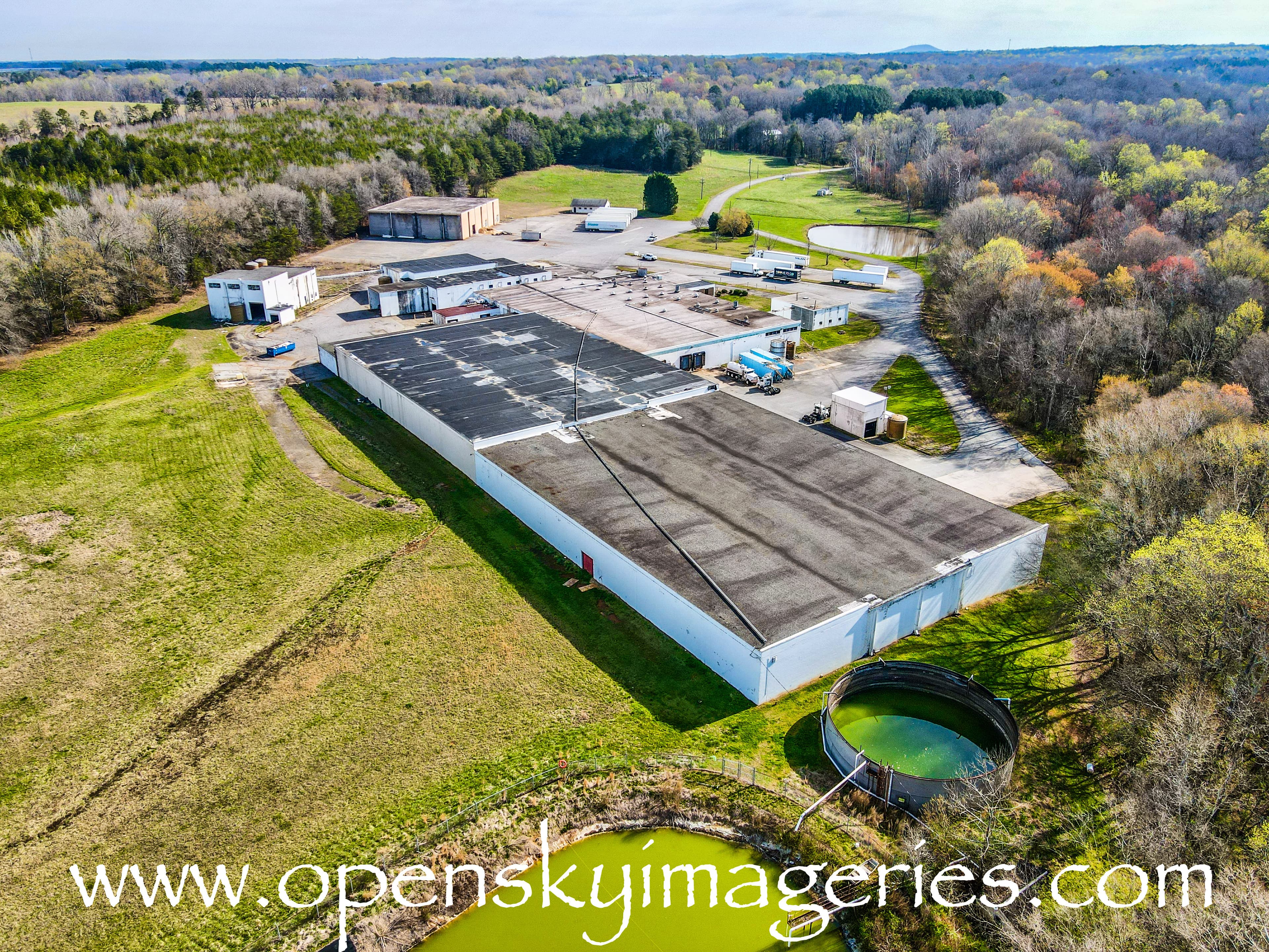Oblique aerial drone photograph of a large industrial facility with connected flat-roof buildings, paved service areas, parked trucks, open grassy land, circular water containment structures, and surrounding woodland.