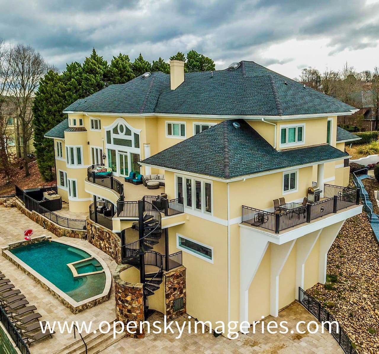 Aerial drone photograph of a large luxury residence with a cream-colored exterior, dark shingle roof, multiple balconies, spiral staircase, stone patio, and private swimming pool with an integrated spa.