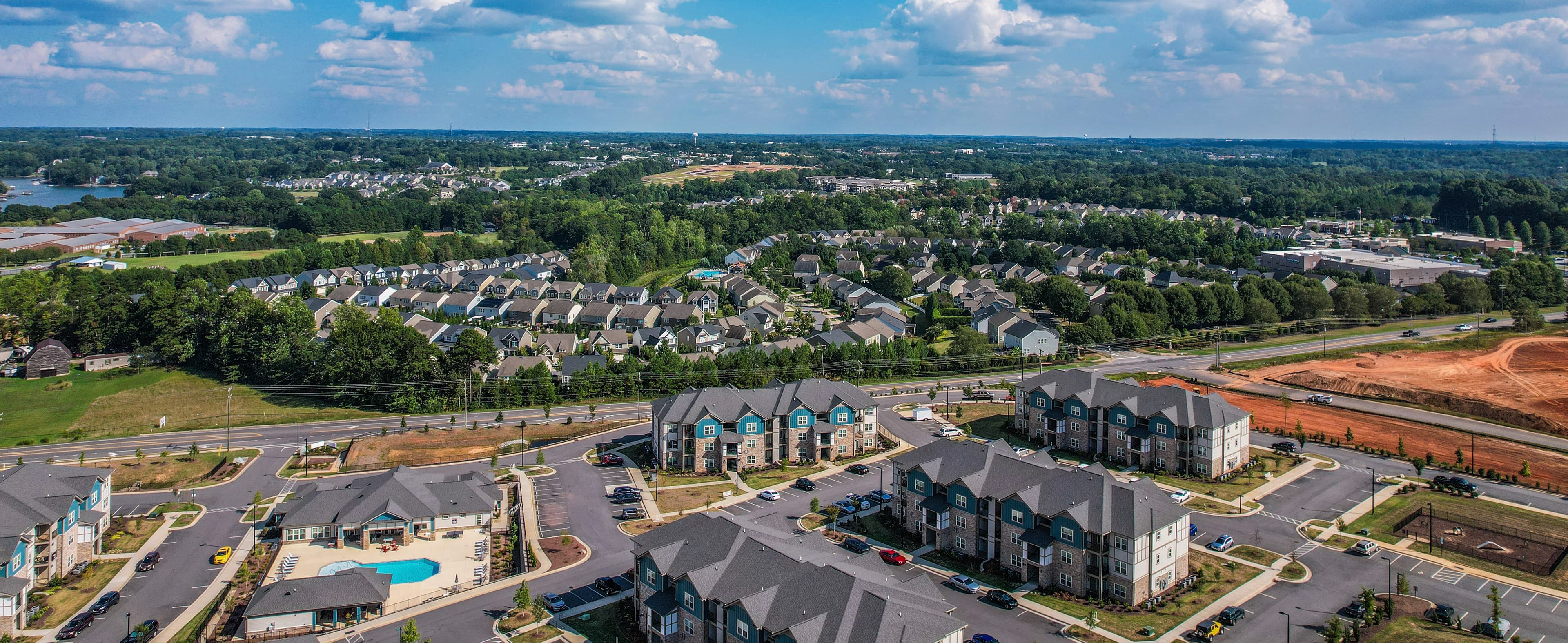 Wide aerial drone photograph of a multifamily apartment community featuring three-story residential buildings, a swimming pool, parking areas, landscaped grounds, and surrounding suburban neighborhoods.