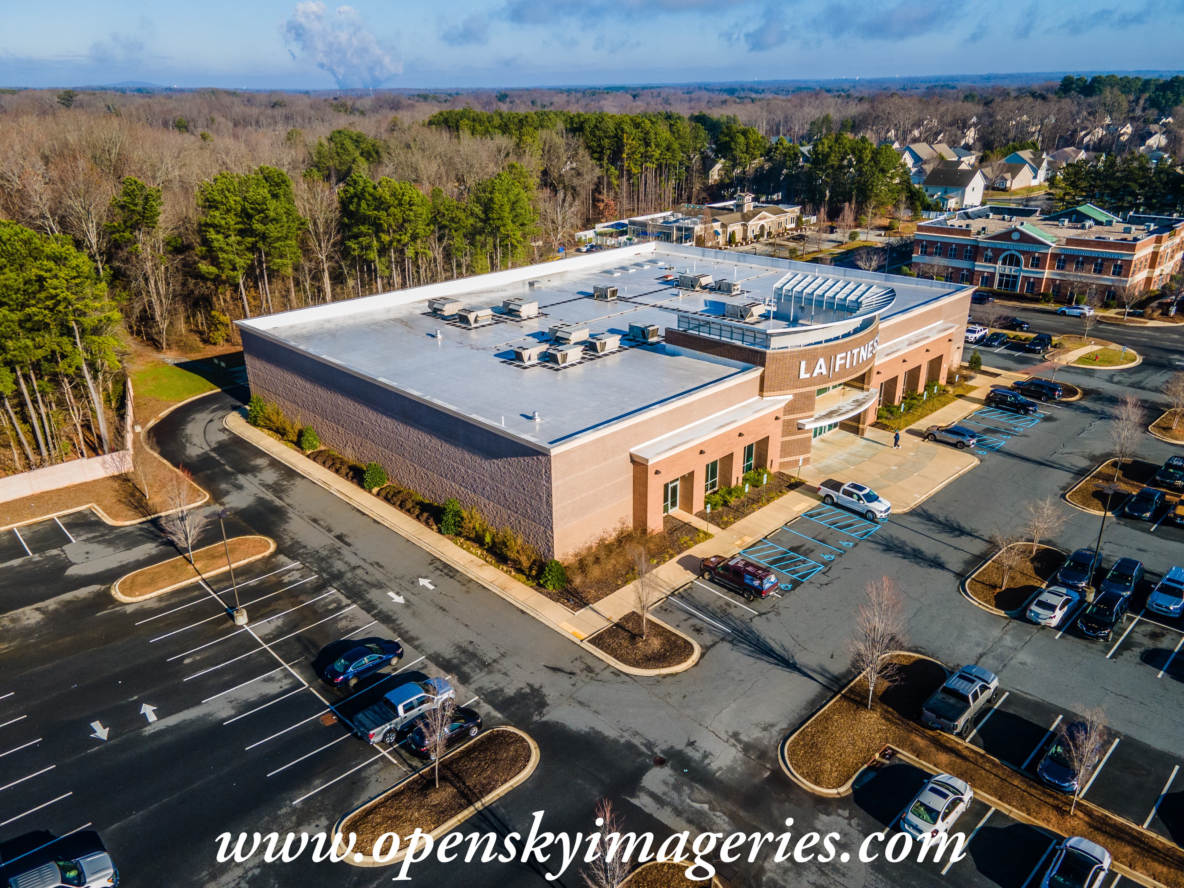 Aerial drone photograph of an LA Fitness gym in a large brick commercial building, showing the flat rooftop, HVAC equipment, main entrance, parking areas, and surrounding businesses and residential neighborhoods.