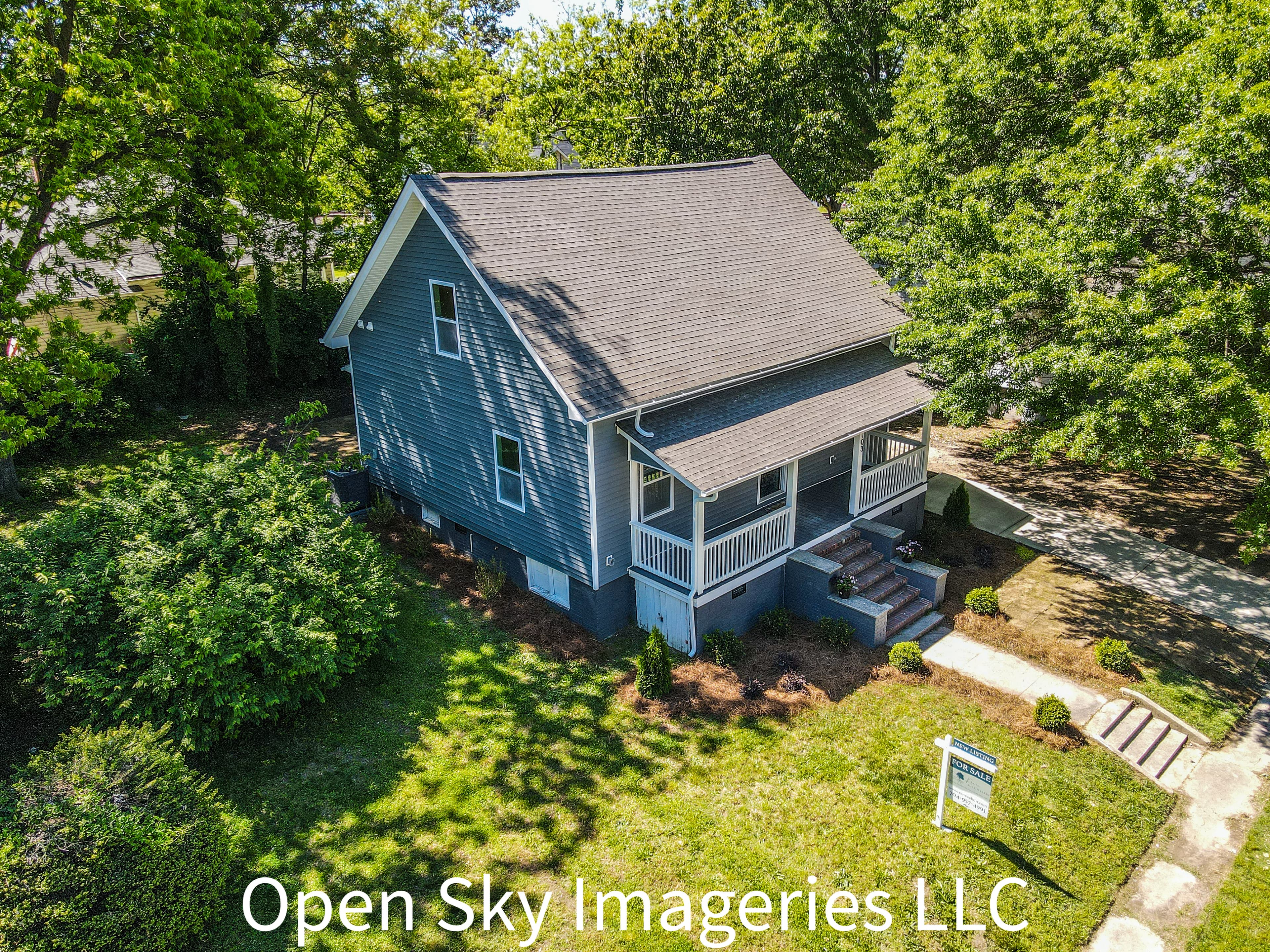 Aerial drone photograph of a two-story gray house for sale, featuring a shingle roof, covered front porch, landscaped lawn, concrete driveway, and mature trees surrounding the property.