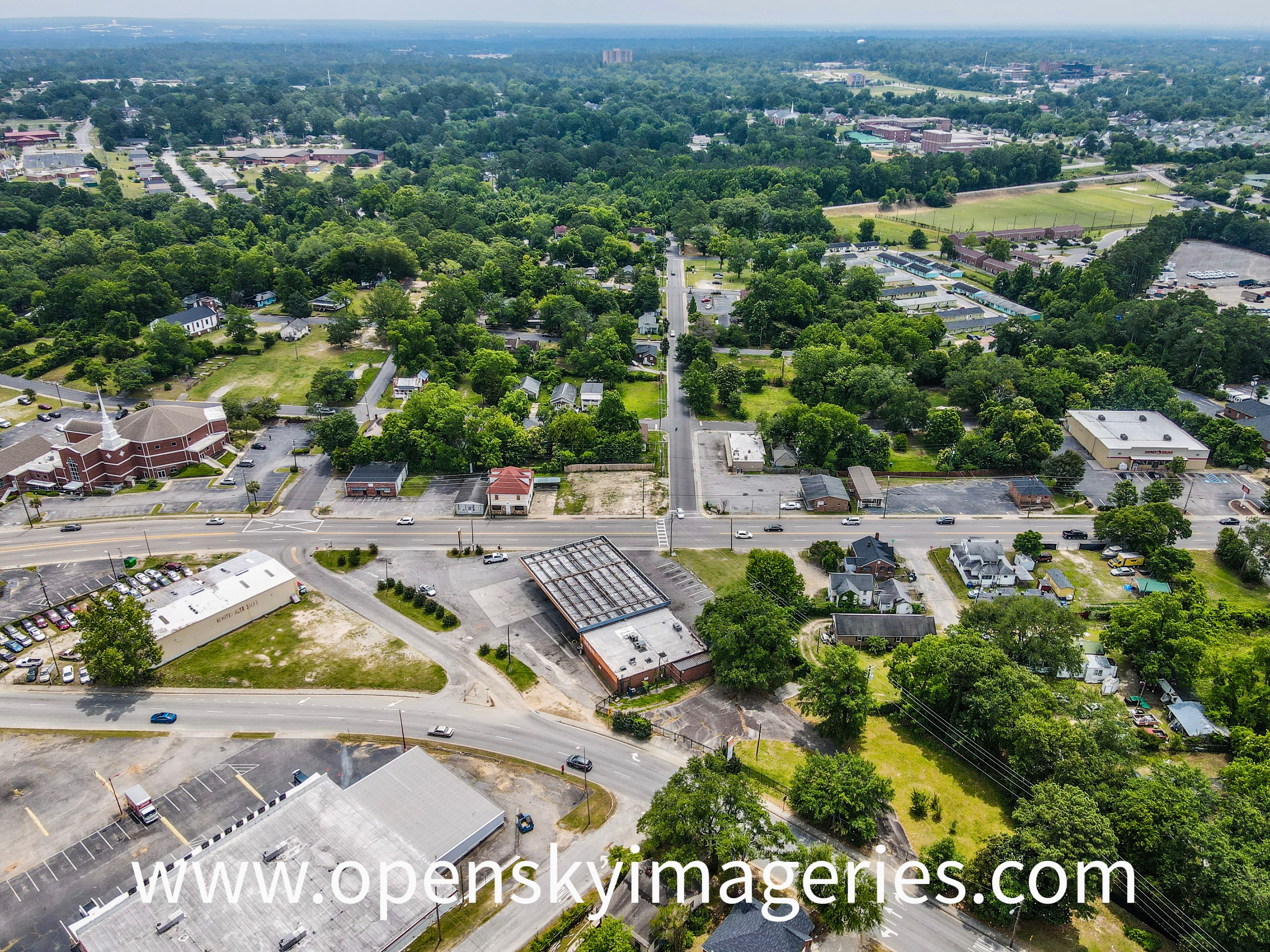 Elevated aerial photograph of a mixed residential and commercial neighborhood, showing a major roadway intersection, retail buildings, parking lots, a church, tree-lined residential streets, and surrounding urban development.