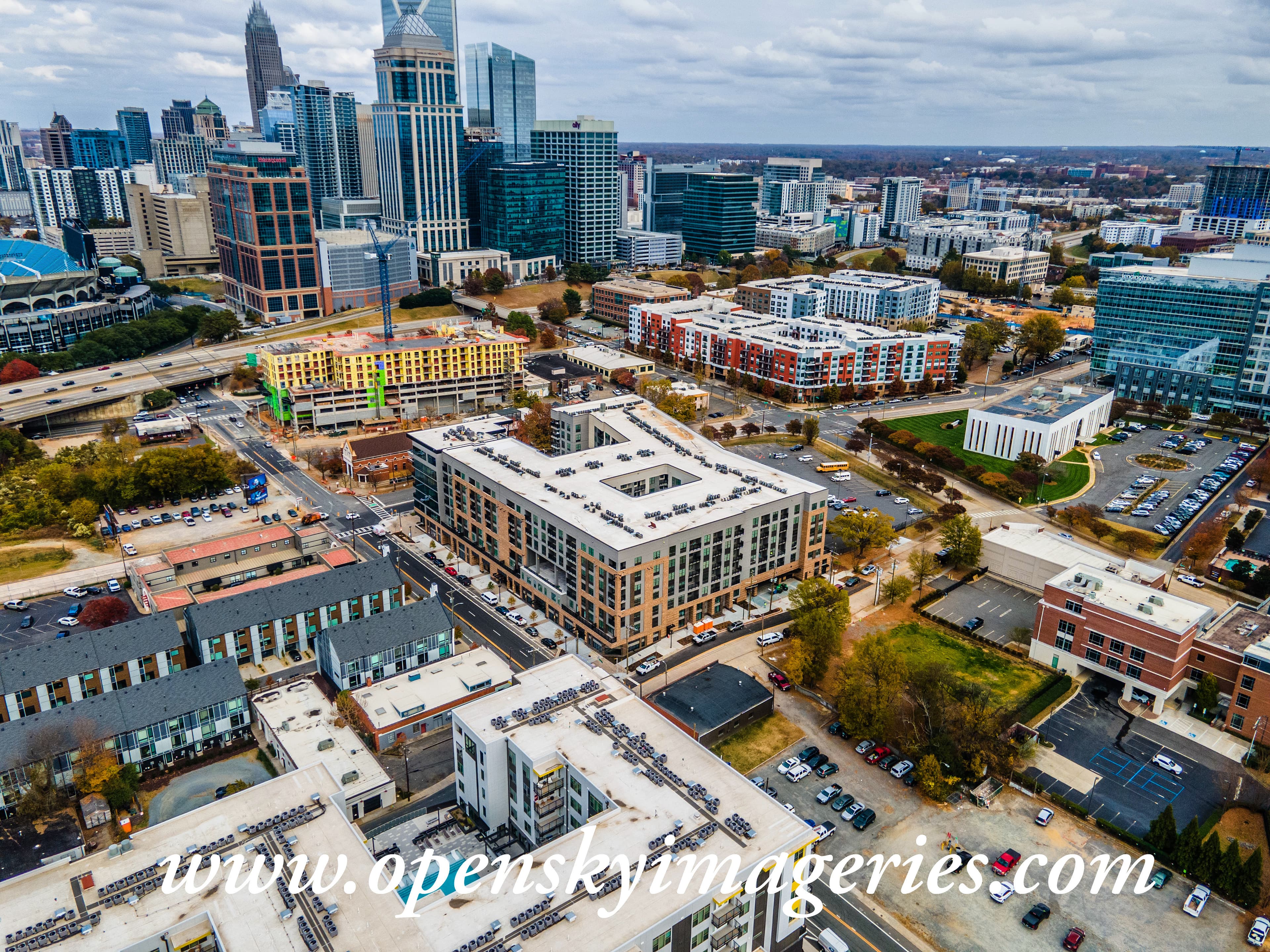 Aerial drone photograph of Uptown Charlotte, North Carolina, showing downtown skyscrapers, mid-rise apartment buildings, commercial properties, an active multistory construction site with a tower crane, and surrounding city streets.